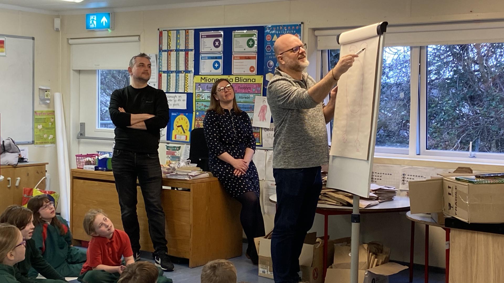 Presenter pointing at a flip chart in a classroom while children sit on the floor listening.