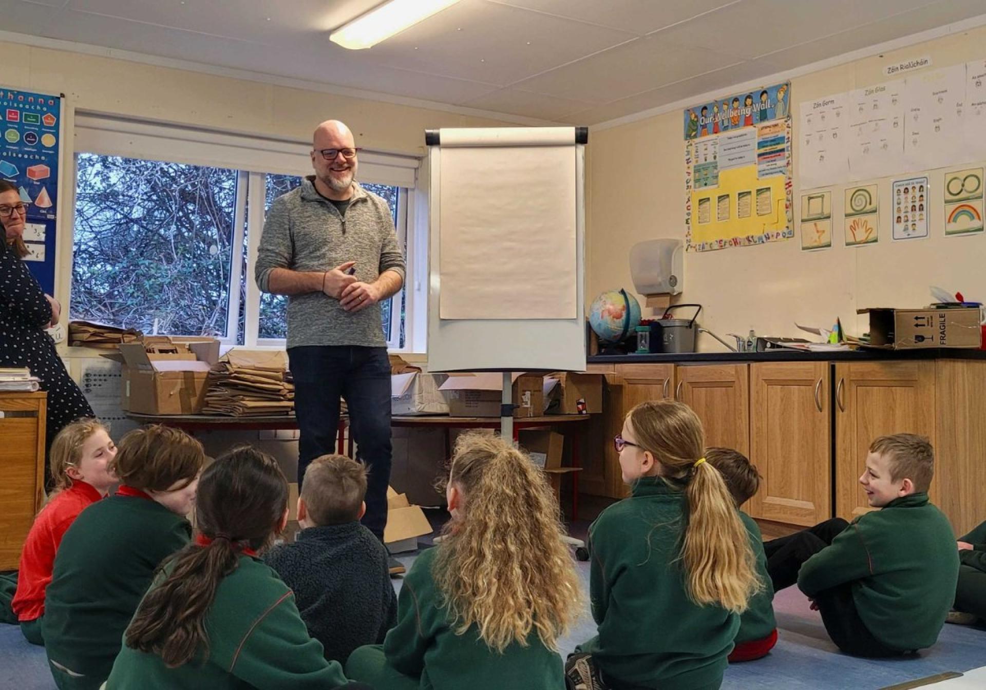 Classroom scene with a presenter standing near a blank flip chart and children seated on the floor.