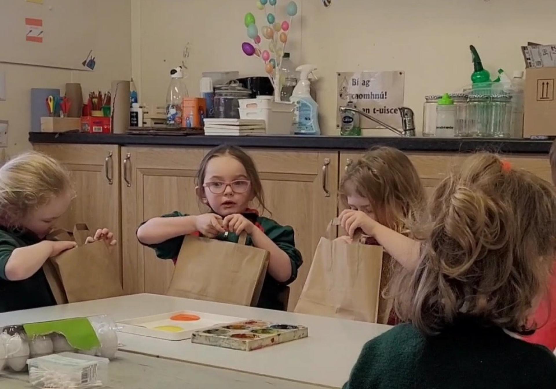 Young children seated at a table opening brown paper Bookbag packages.
