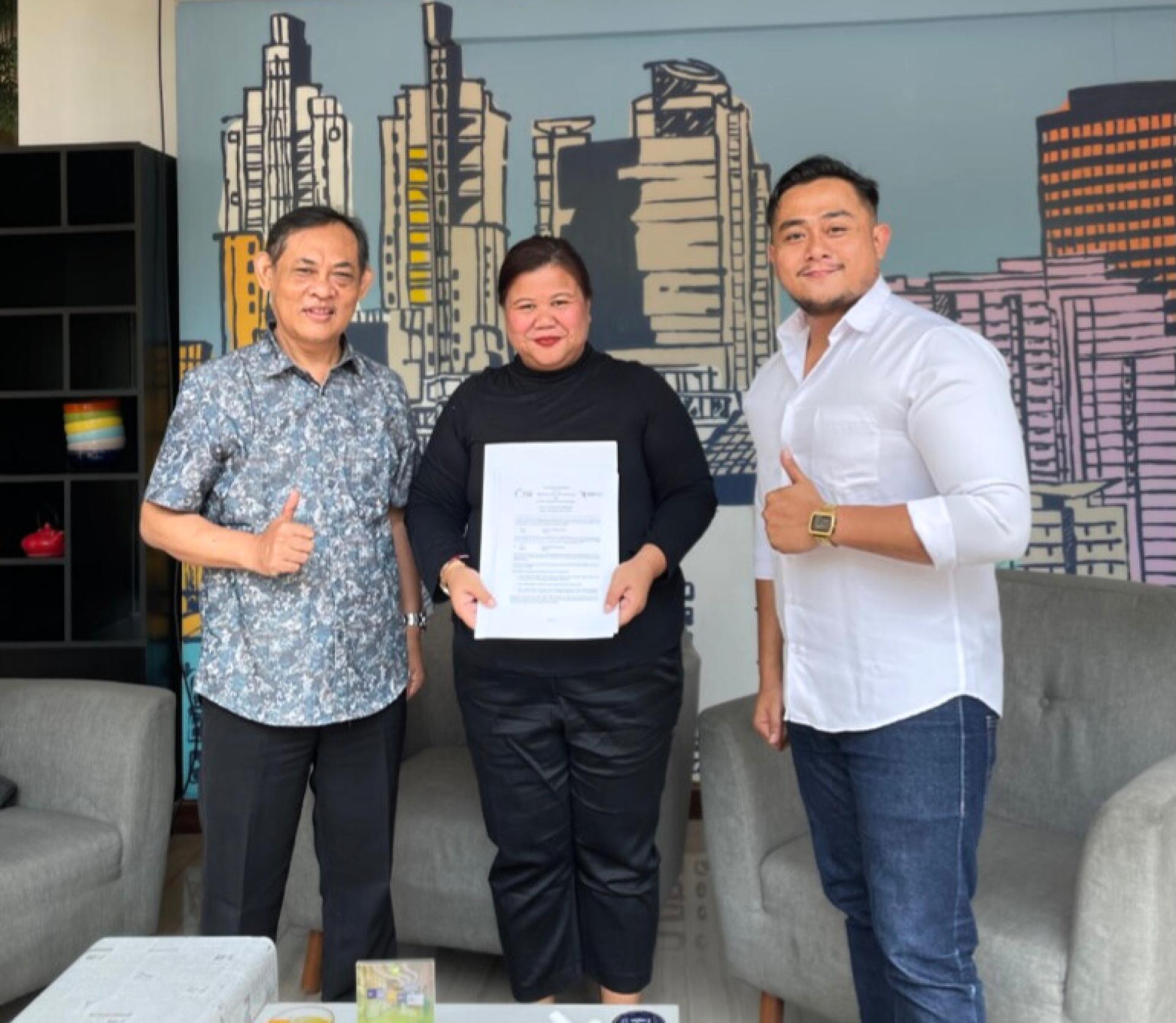 Three people standing indoors, one holding a signed document while others give thumbs-up gestures.
