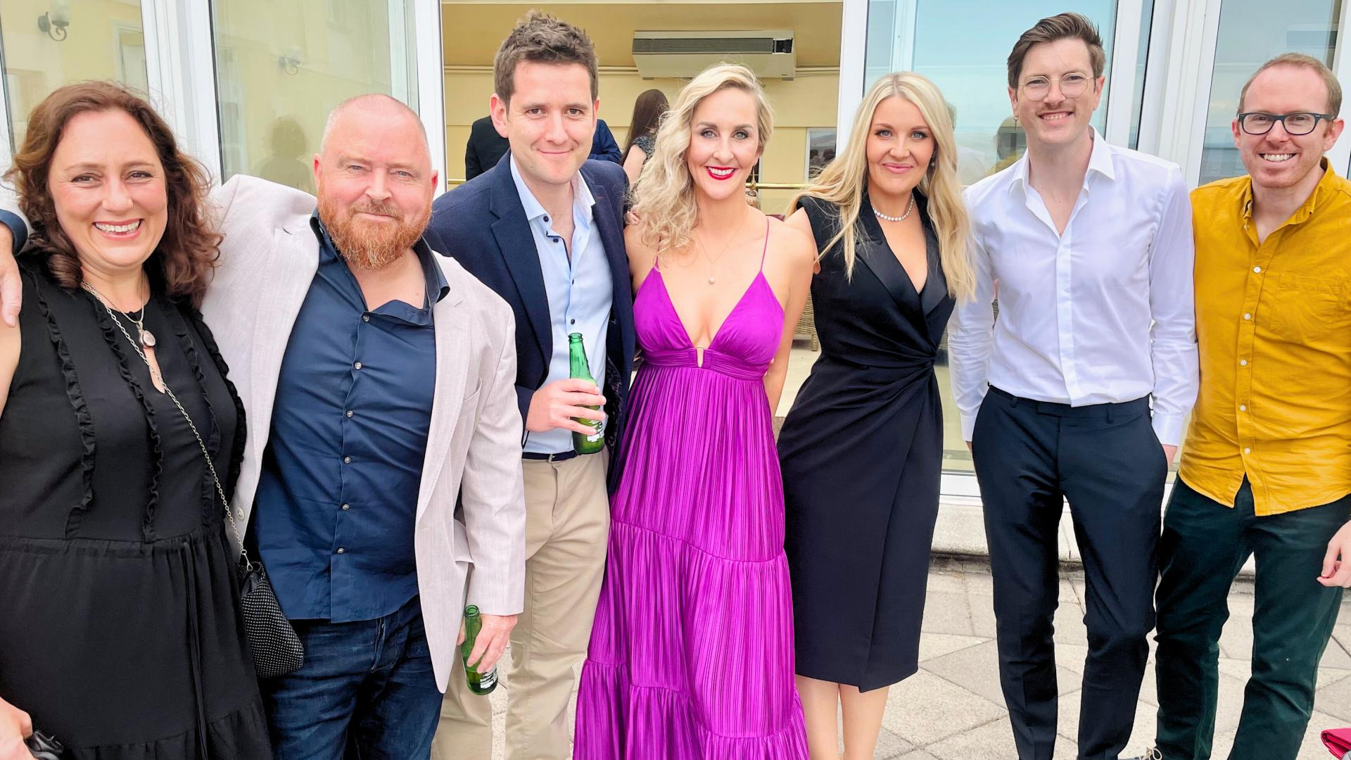 Group of people posing outdoors, some holding drinks, dressed in formal and semi-formal attire.