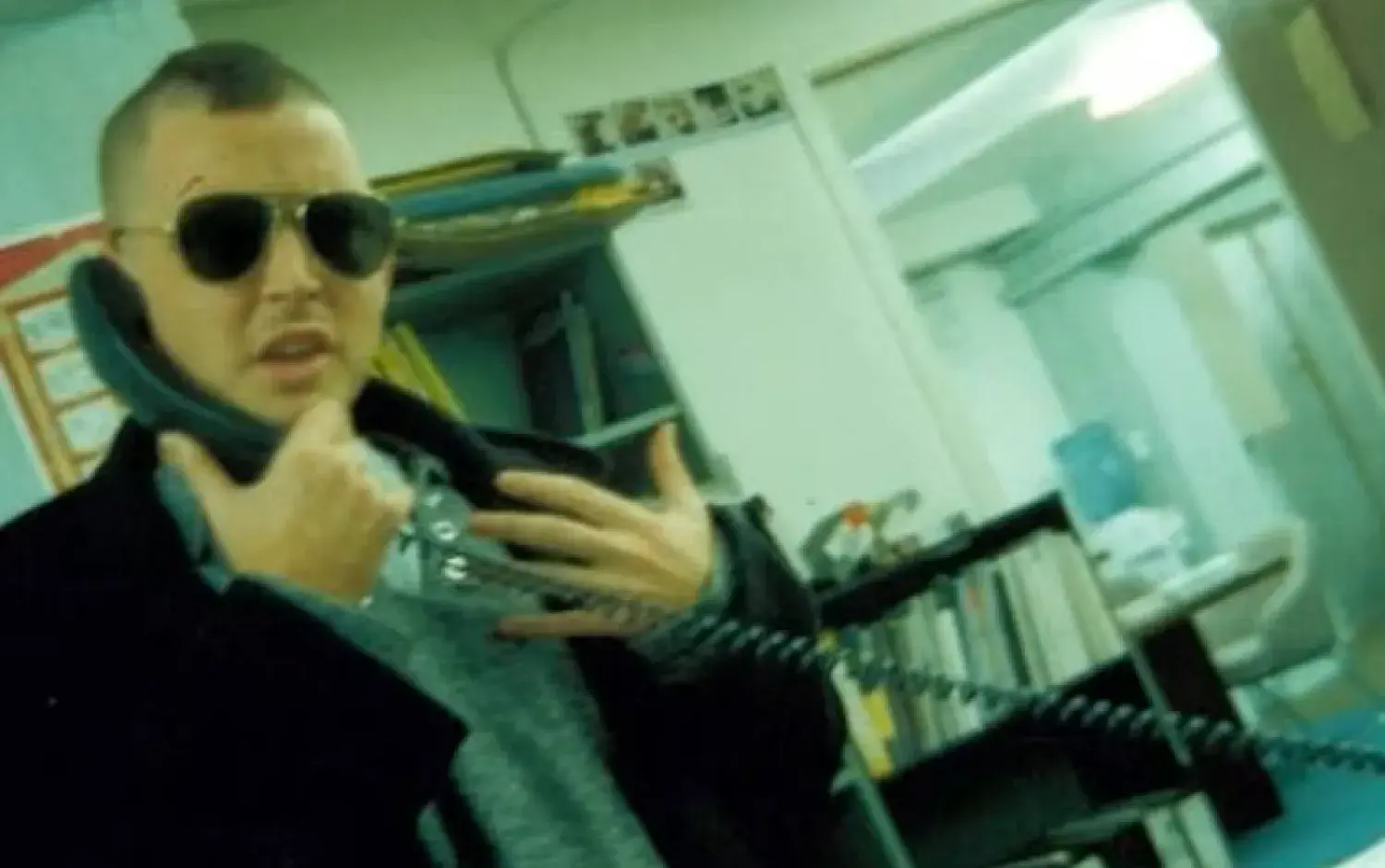 A person wearing sunglasses holds a phone receiver to their ear in an office setting with shelves of books and binders in the background.