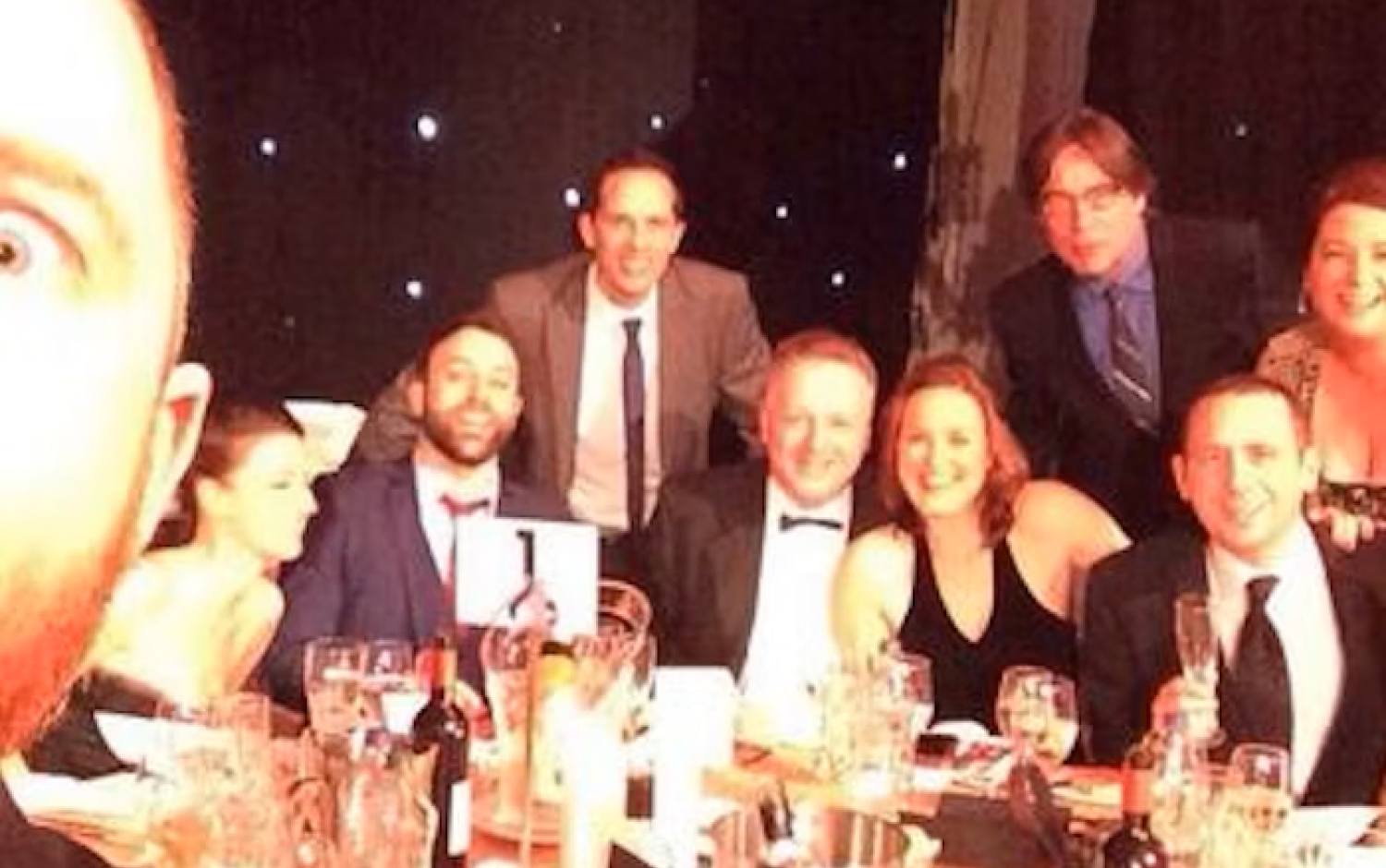 A group of people in formal attire seated at a table set for dining, with one person taking a selfie in front of a starry curtain backdrop.
