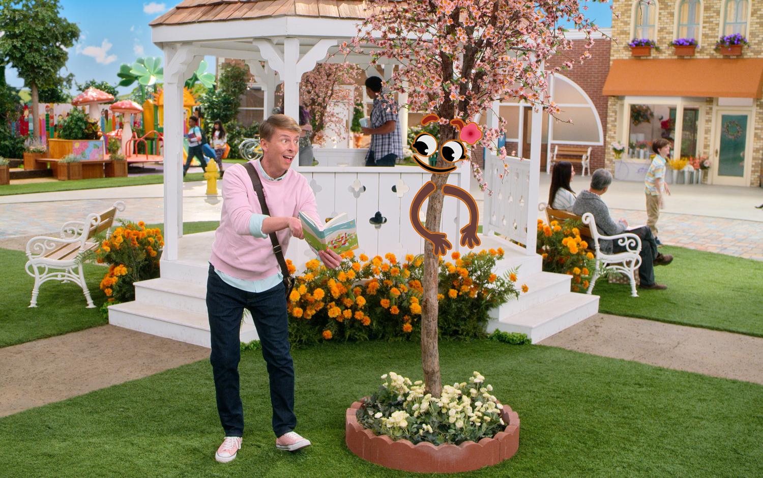Colorful town square with a white gazebo, blooming flowers, and a playful cartoon-faced tree with a pink bow; person reading a book nearby in a vibrant outdoor setting.