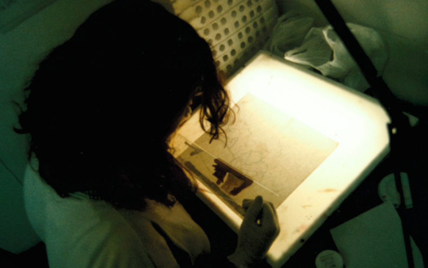 Person working at a lightbox, tracing or painting animation cels in a dimly lit studio.