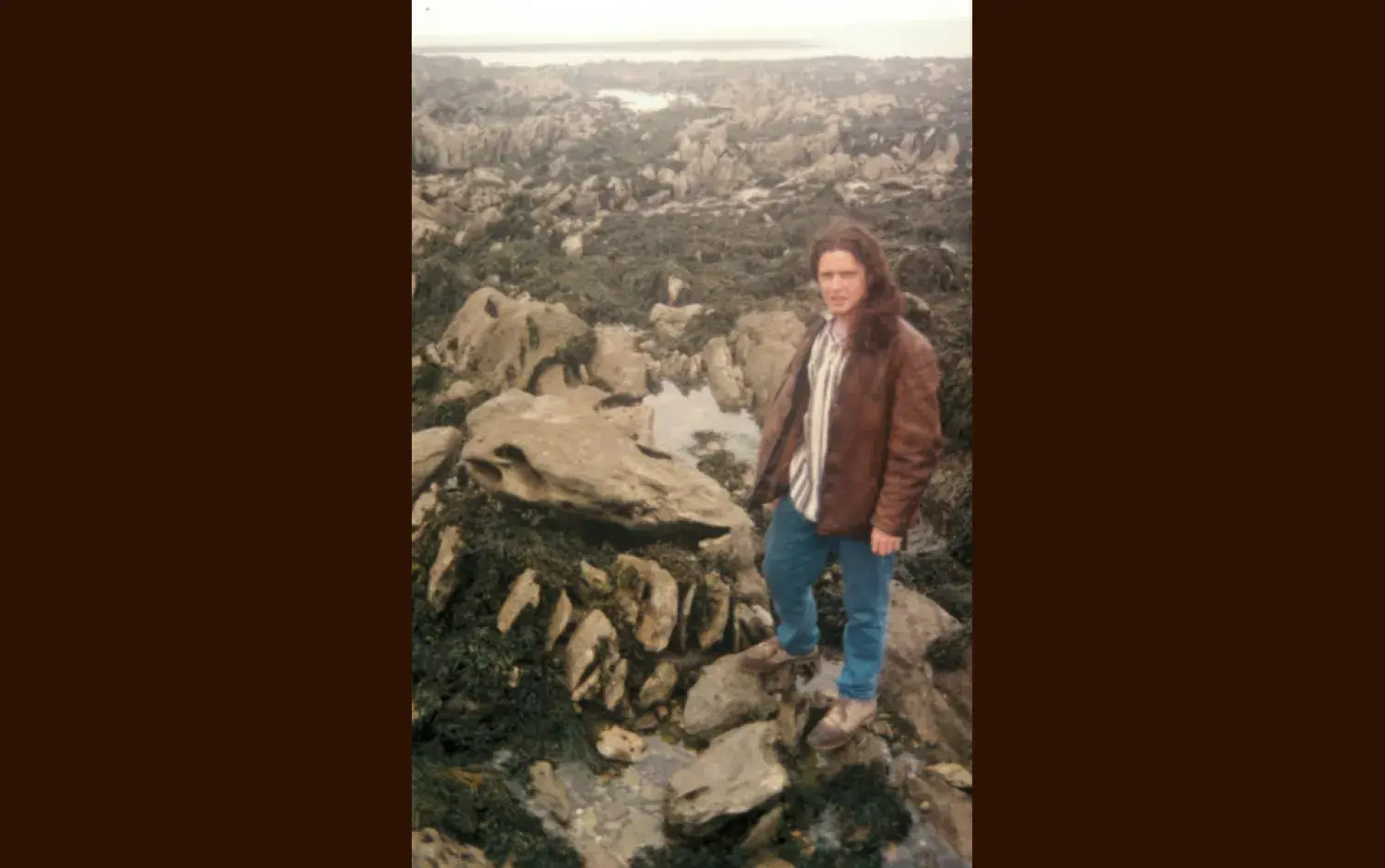 Individual standing on rocky, seaweed-covered shoreline with tidal pools in the background.