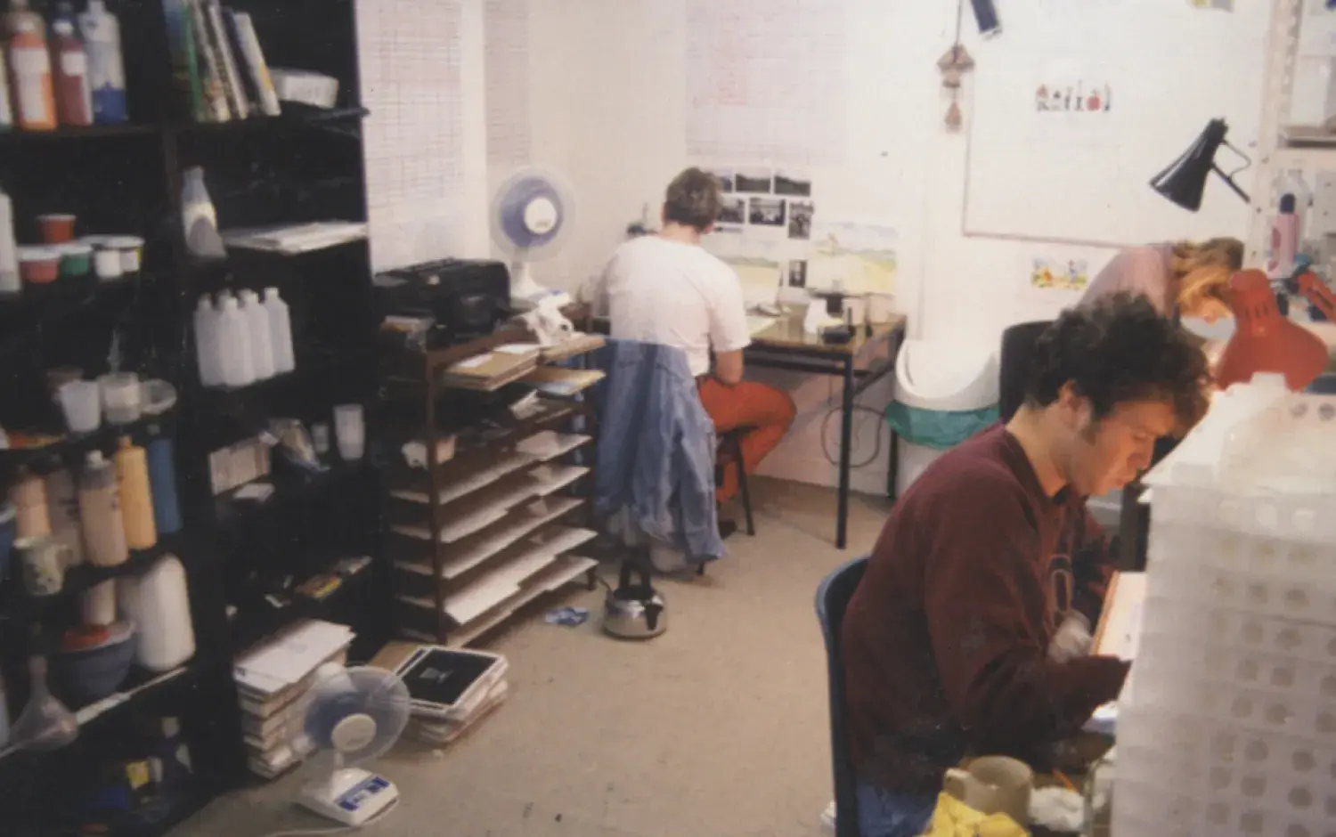 Animation studio workspace with shelves of supplies and three people seated at desks working on artwork.