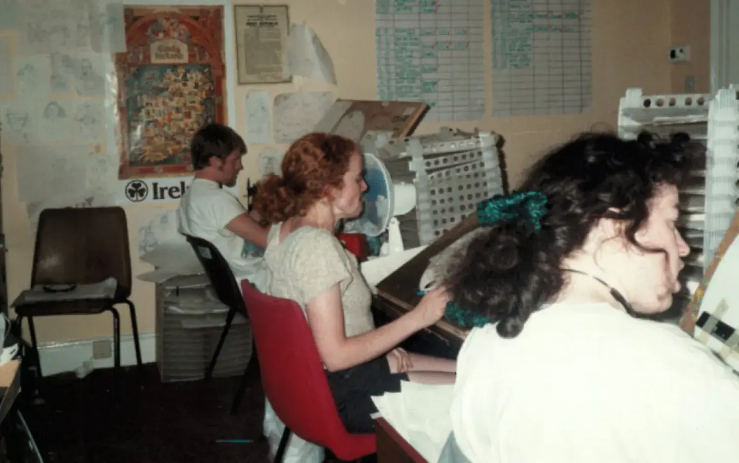 Three animators seated at desks painting animation cels, surrounded by shelves and pinned artwork.