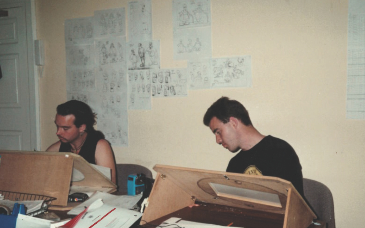 Two animators working at wooden animation desks with character sketches pinned to the wall behind them.
