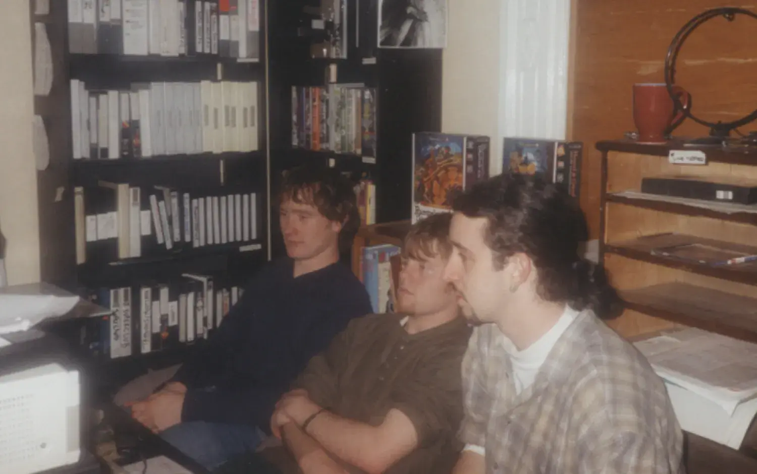 Three individuals seated in front of shelves filled with VHS tapes and books in a small office space.