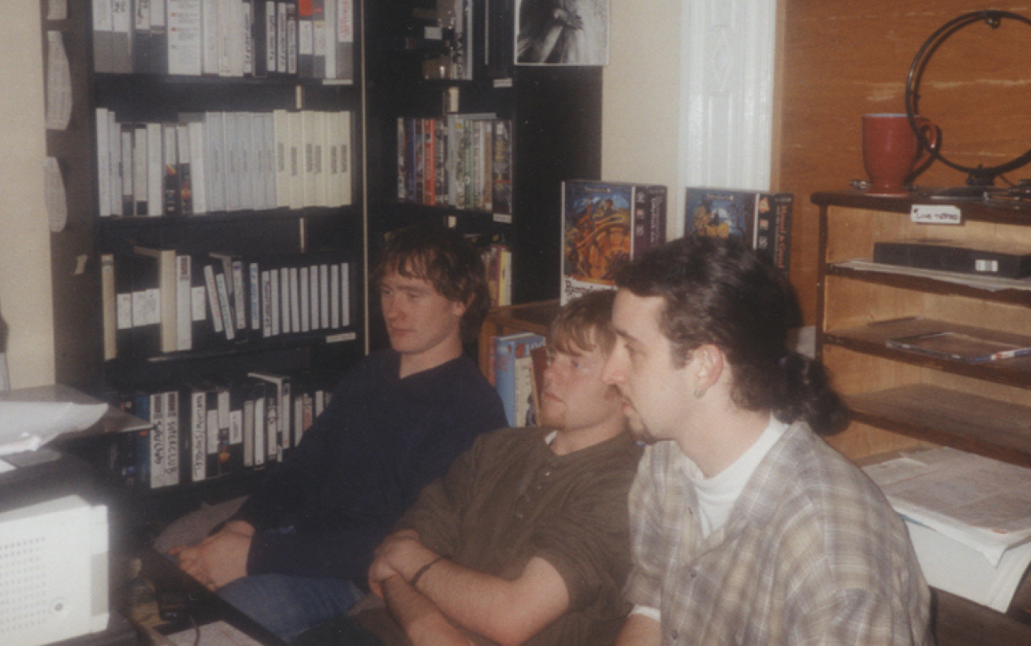 Three individuals seated in front of shelves filled with VHS tapes and books in a small office space.