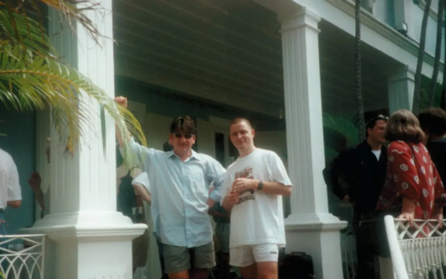 Group of people gathered on a porch with white columns and tropical plants, two individuals standing in the foreground.