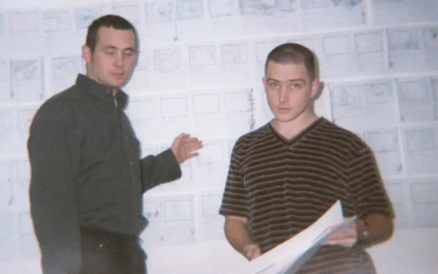 Two people standing in front of a wall covered with storyboard sketches, one holding a large sheet of paper.
