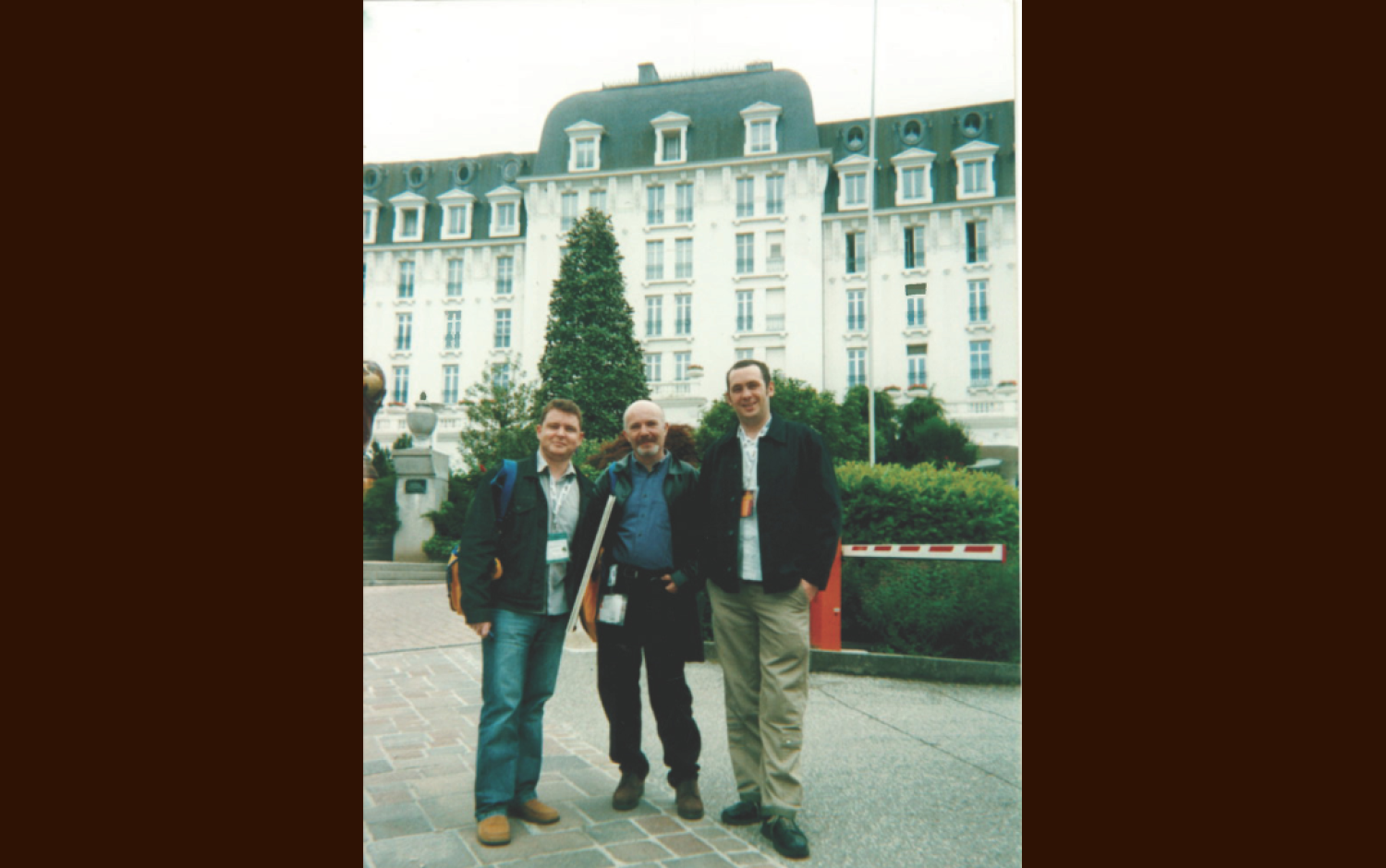 Three people standing outdoors in front of a large, ornate building with a mansard roof and landscaped grounds.
