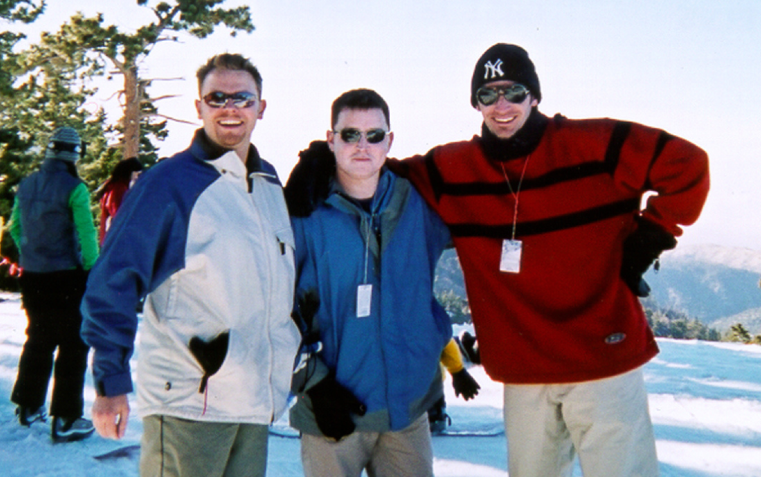 Three people standing on a snowy slope with ski gear, trees, and mountains in the background.