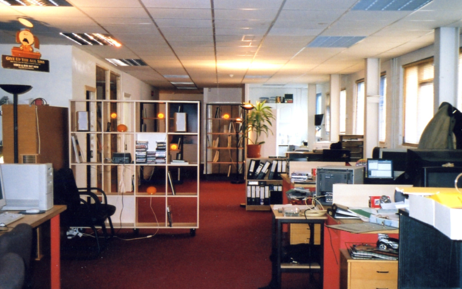 Wide view of an open-plan office with desks, computers, shelving units, and a red carpeted floor.