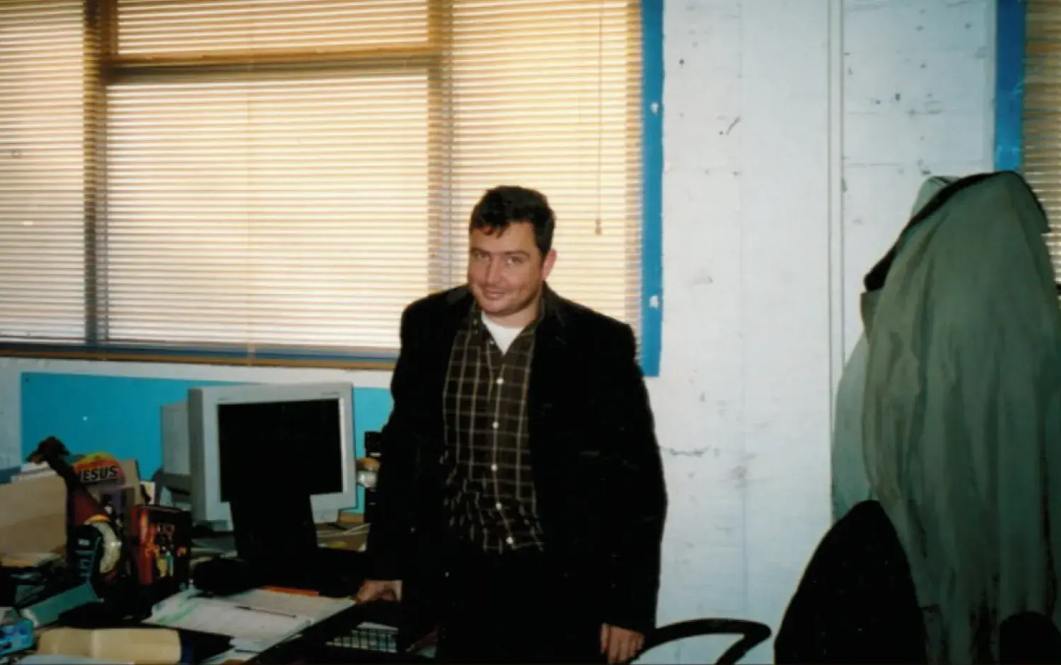 Office workspace with a desk, computer monitor, and a coat hanging on the wall; person standing near the desk.