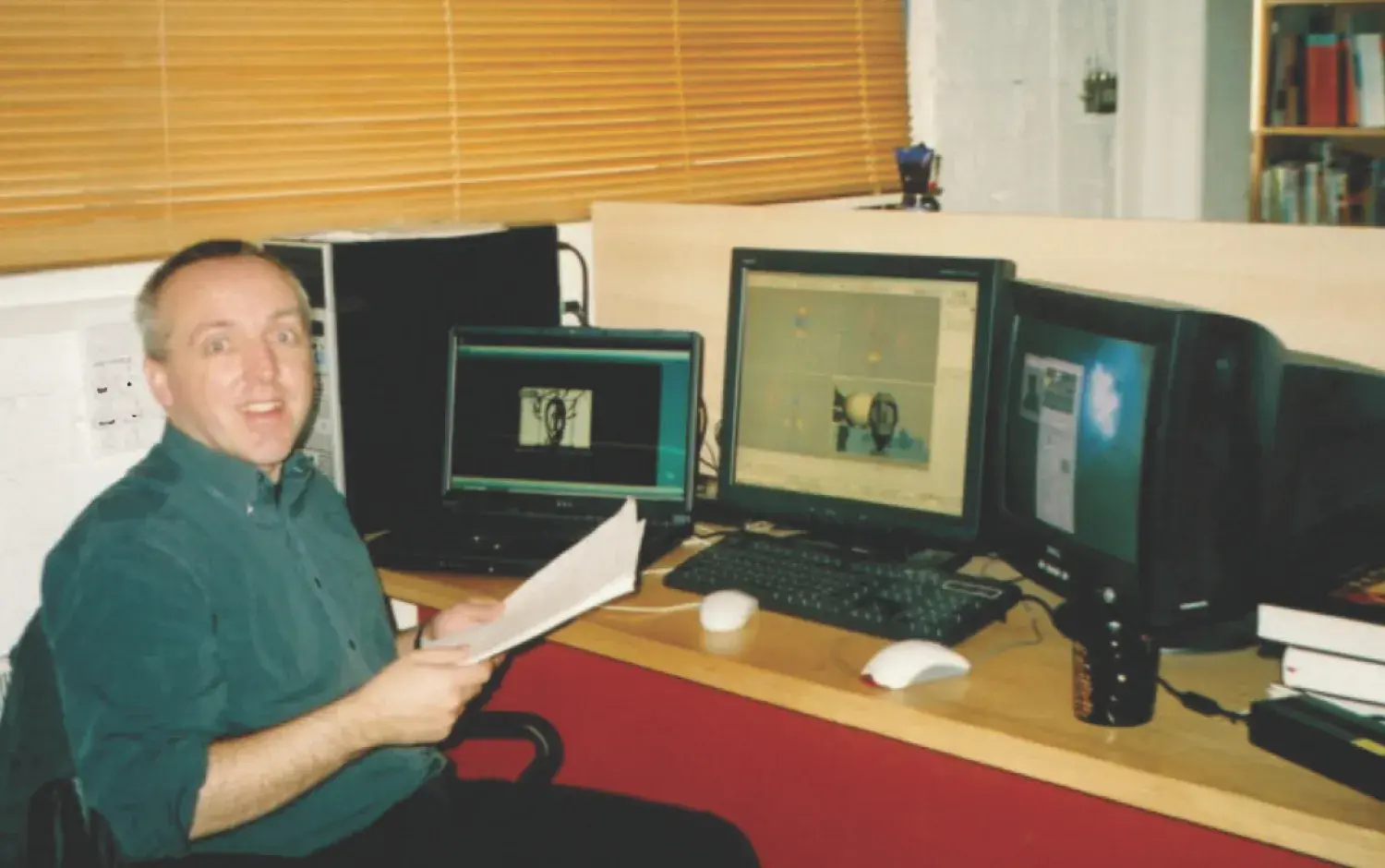 Person at a workstation with multiple monitors showing animation software, holding a sheet of paper.