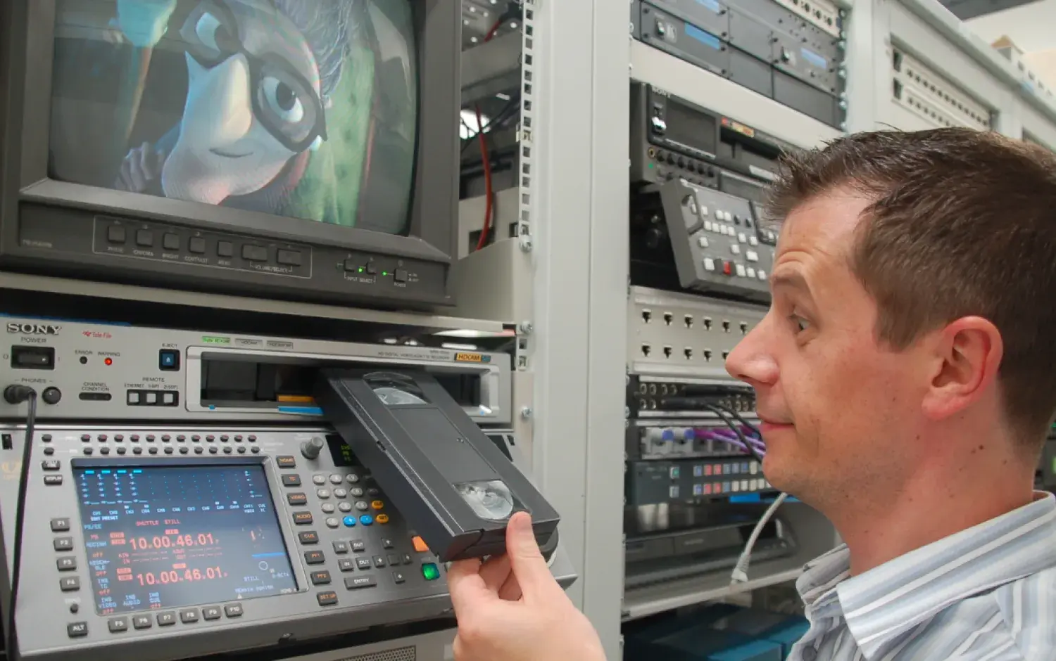 Person inserting a VHS tape into a professional video deck; monitor above shows a 3D animated character.