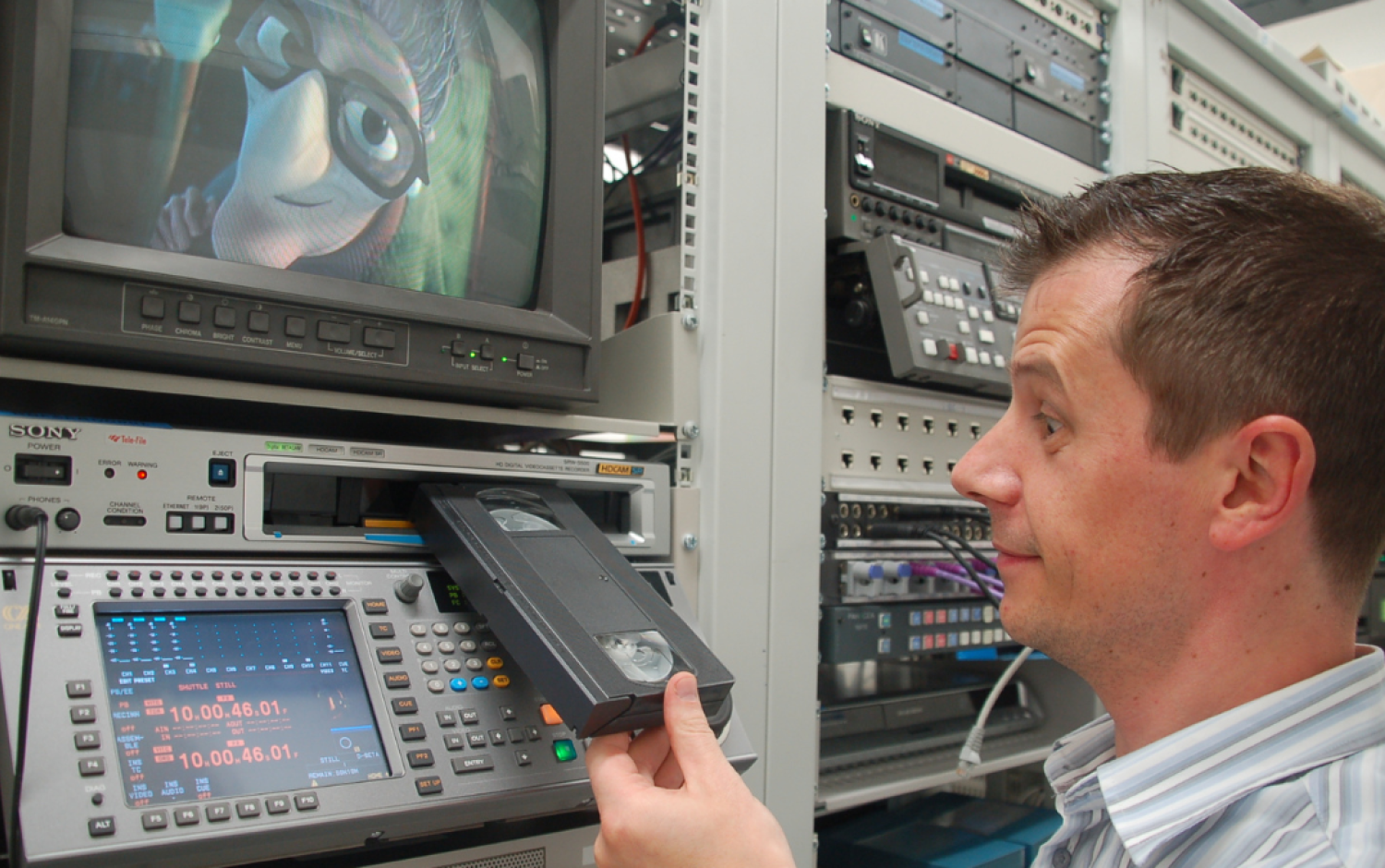 Person inserting a VHS tape into a professional video deck; monitor above shows a 3D animated character.