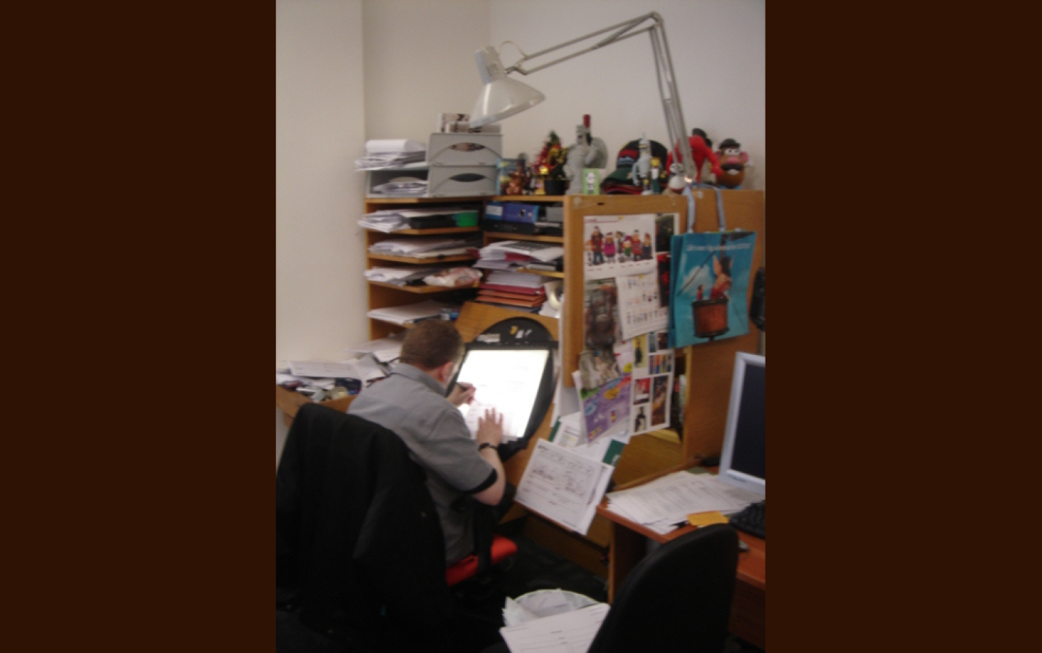 Animator seated at a cluttered desk using a lightbox to work on hand-drawn animation sheets; shelves behind are filled with papers and figurines.