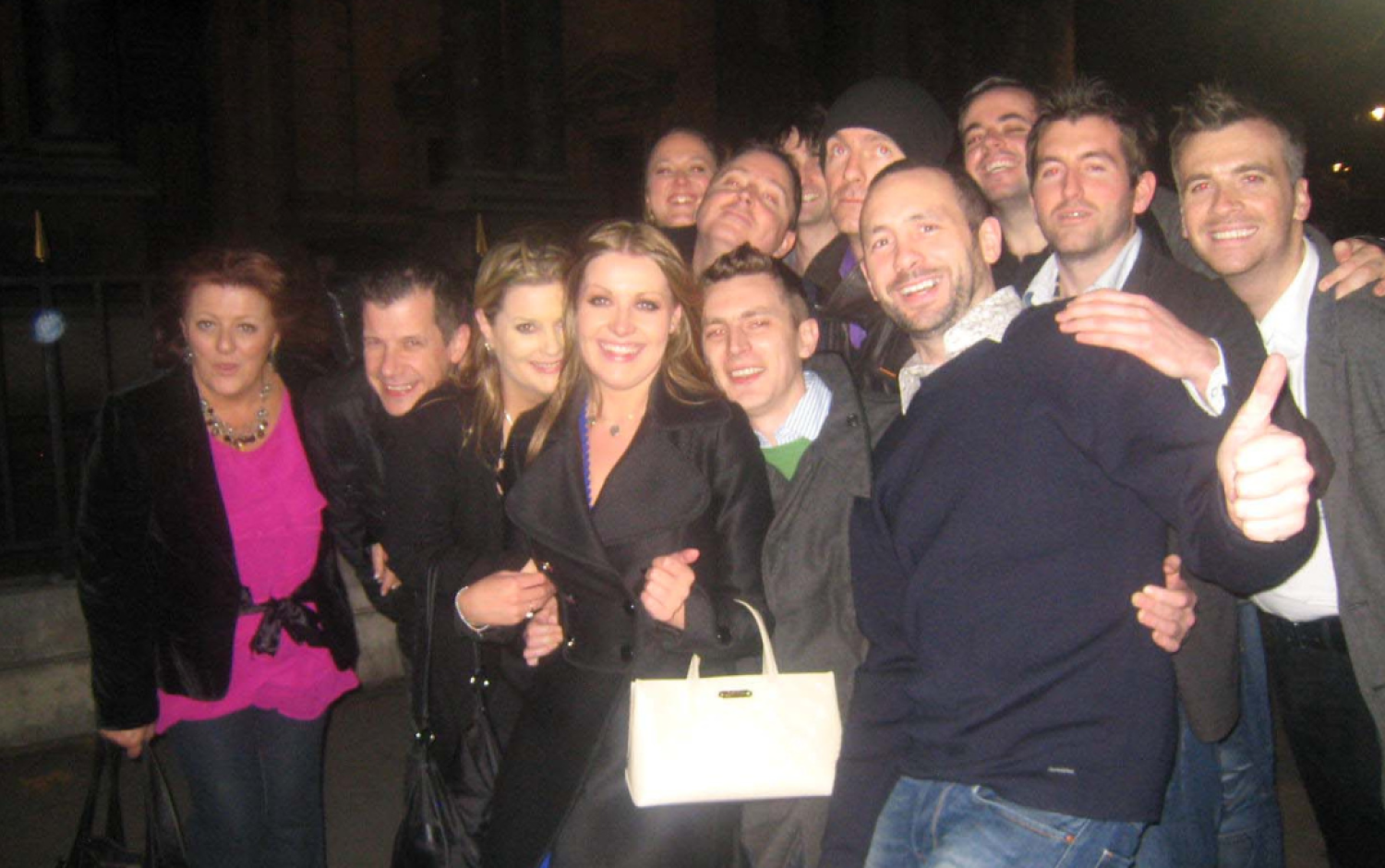 Group of people posing closely together outdoors at night, some with arms around each other.