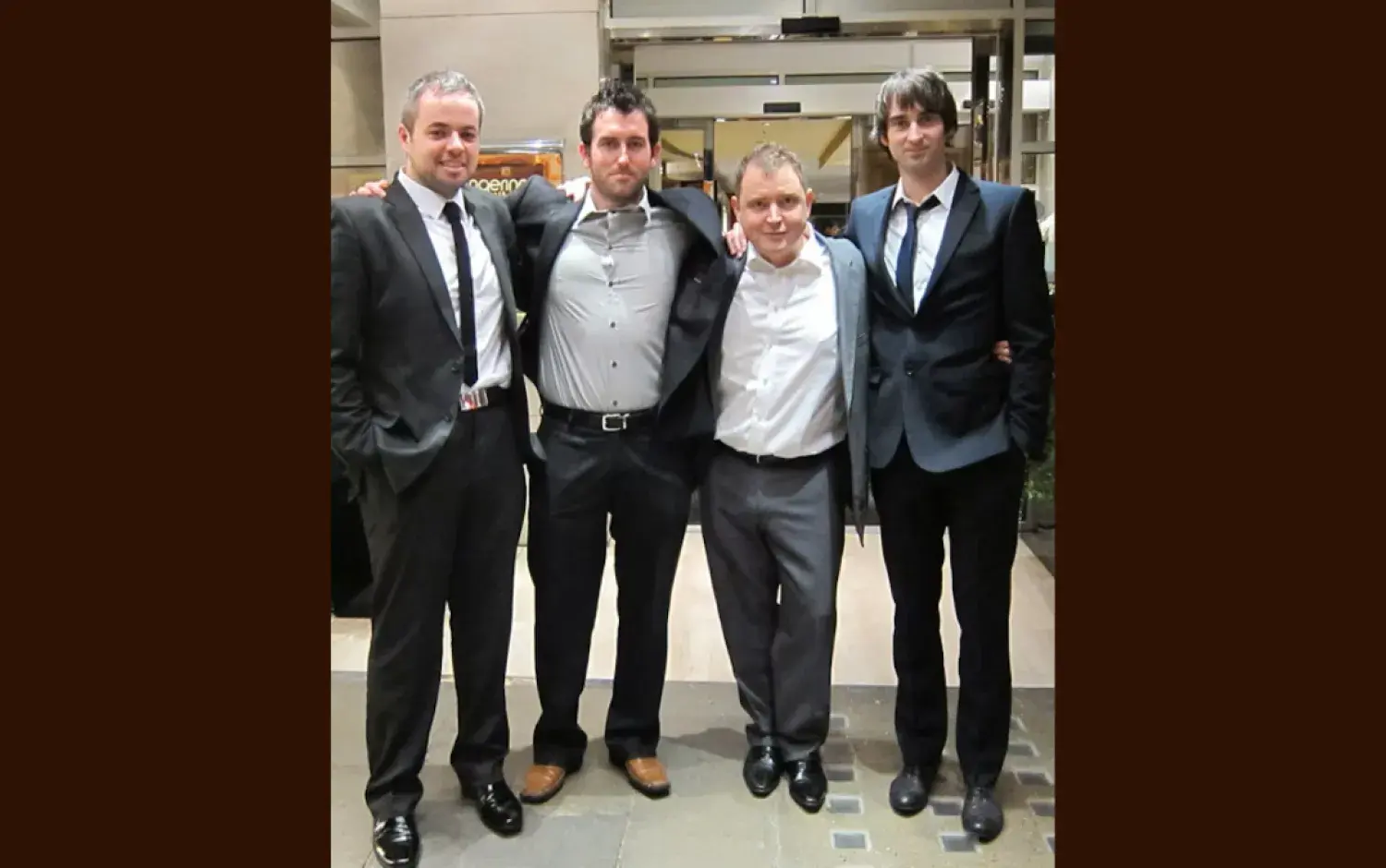 Four people dressed in formal suits standing together in a hotel lobby, arms linked.