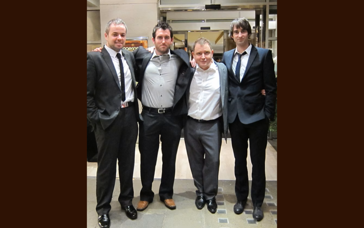 Four people dressed in formal suits standing together in a hotel lobby, arms linked.