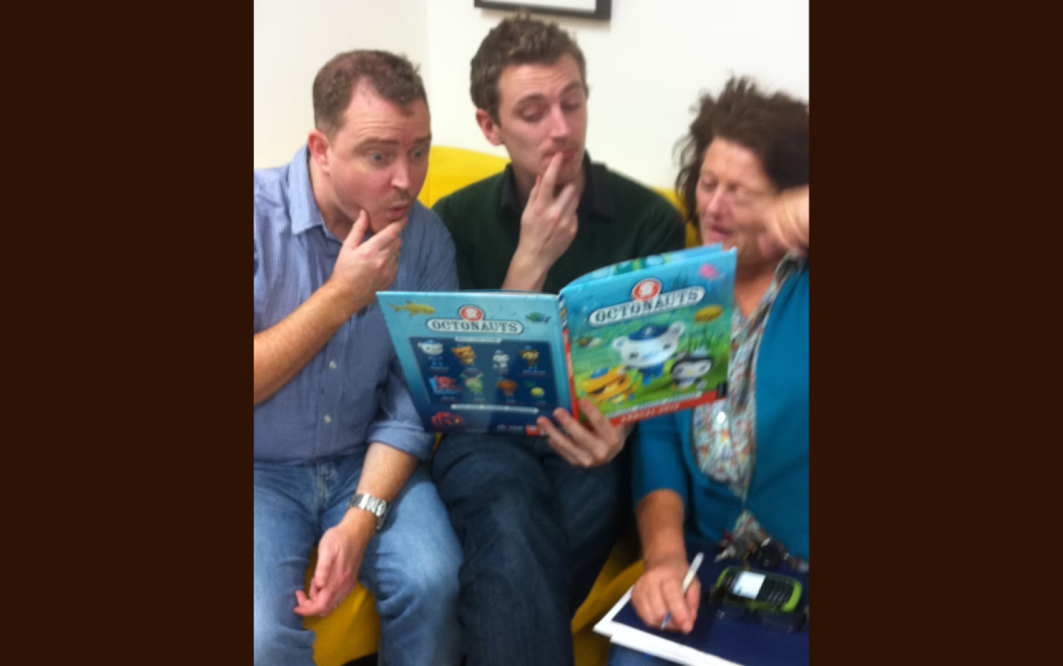 Three people sitting on a yellow couch reading an Octonauts book, with another person holding a notepad.