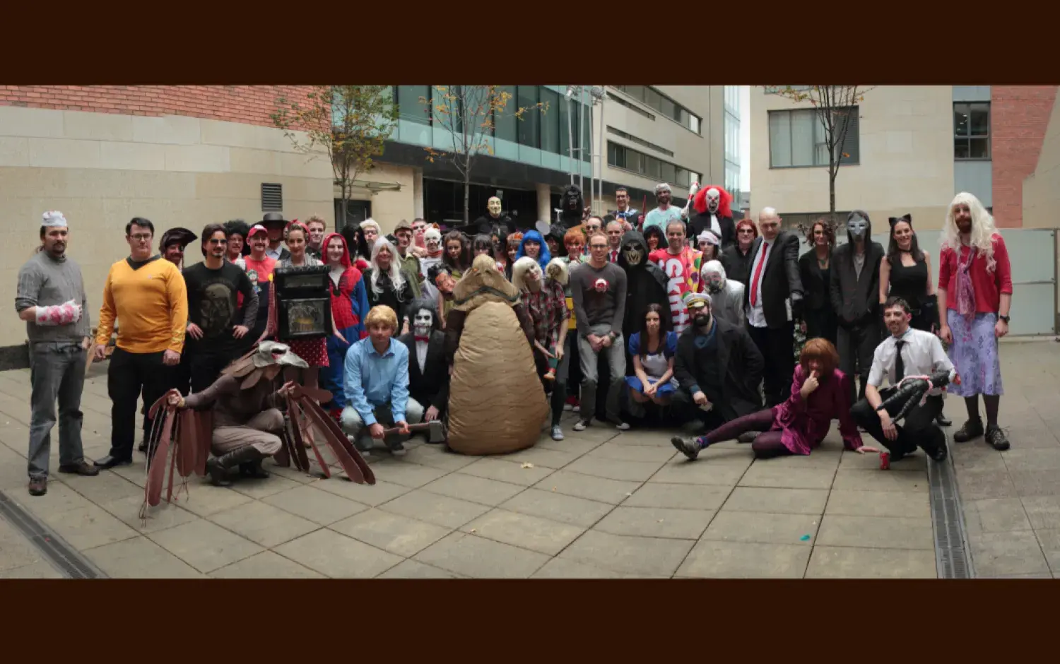 Large group of people in costumes gathered in a courtyard for a Halloween event, featuring various props and themed outfits.