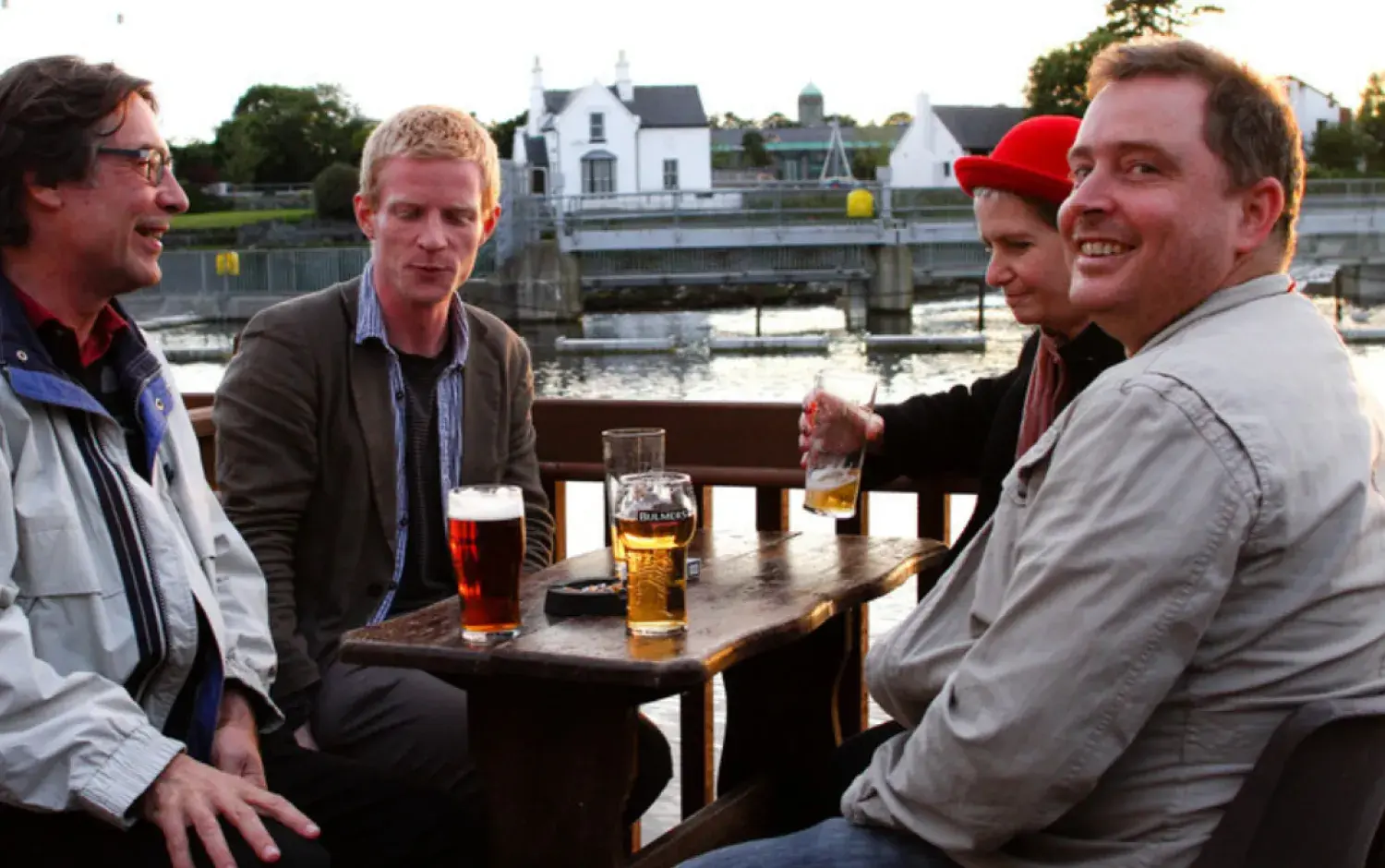 Four people sitting at an outdoor wooden table with glasses of beer and cider, overlooking a river and white buildings in the background.