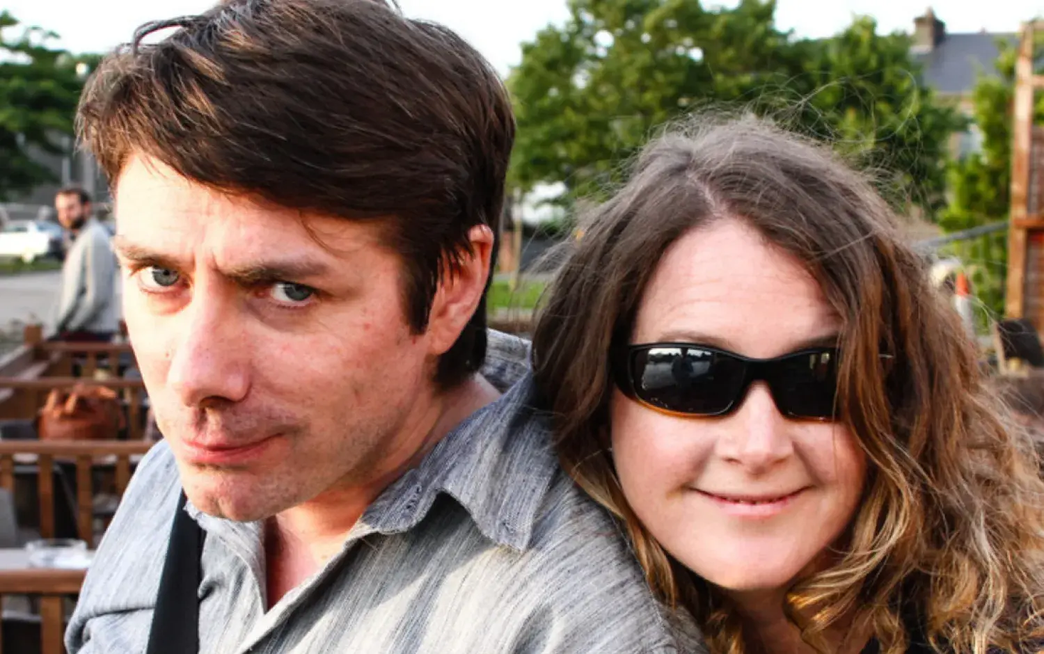 Two people sitting close together outdoors at a wooden table in a casual setting, with empty chairs and greenery visible in the background.