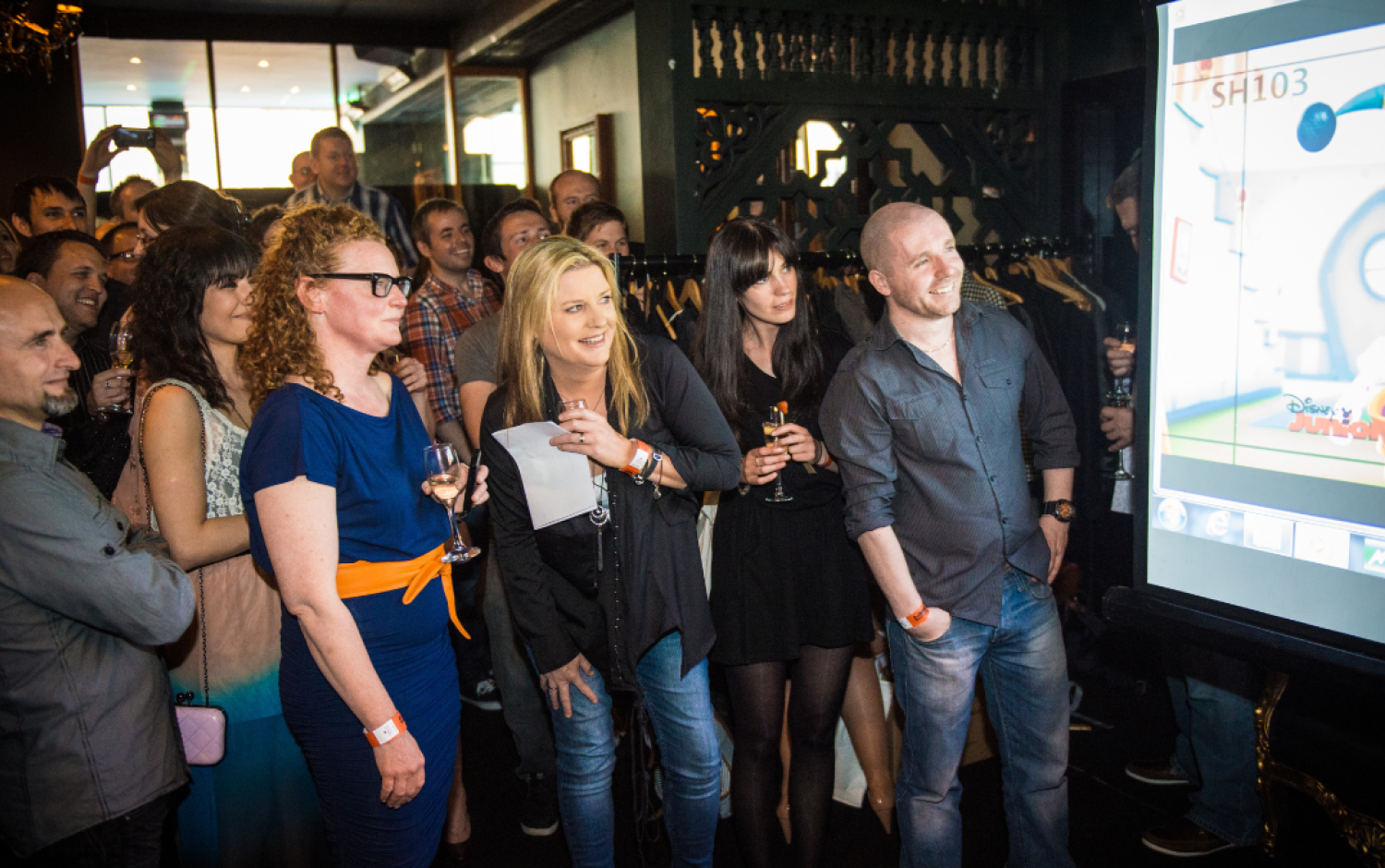 Crowd gathered in a dimly lit venue watching an animated character projected on a large screen; some people holding drinks.