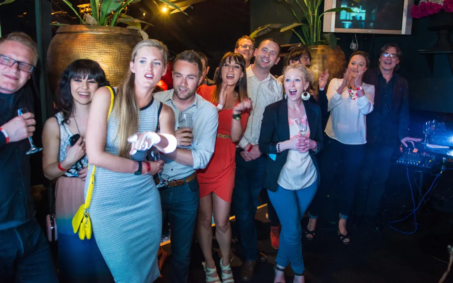 Crowd standing in a bar area, holding drinks, with large decorative vases and plants in the background.