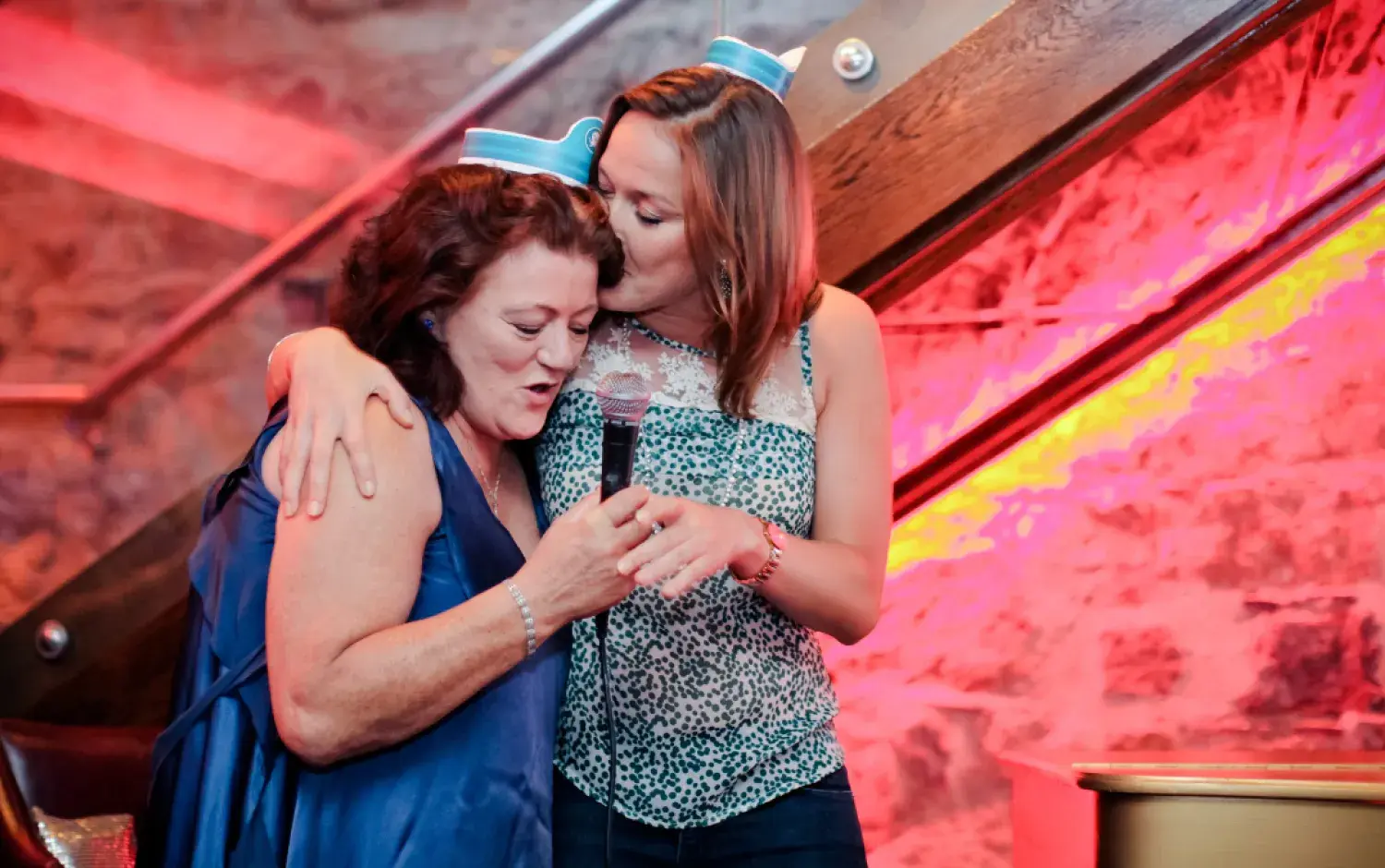 Two people embracing while holding a microphone on a stage with red accent lighting and stone walls.