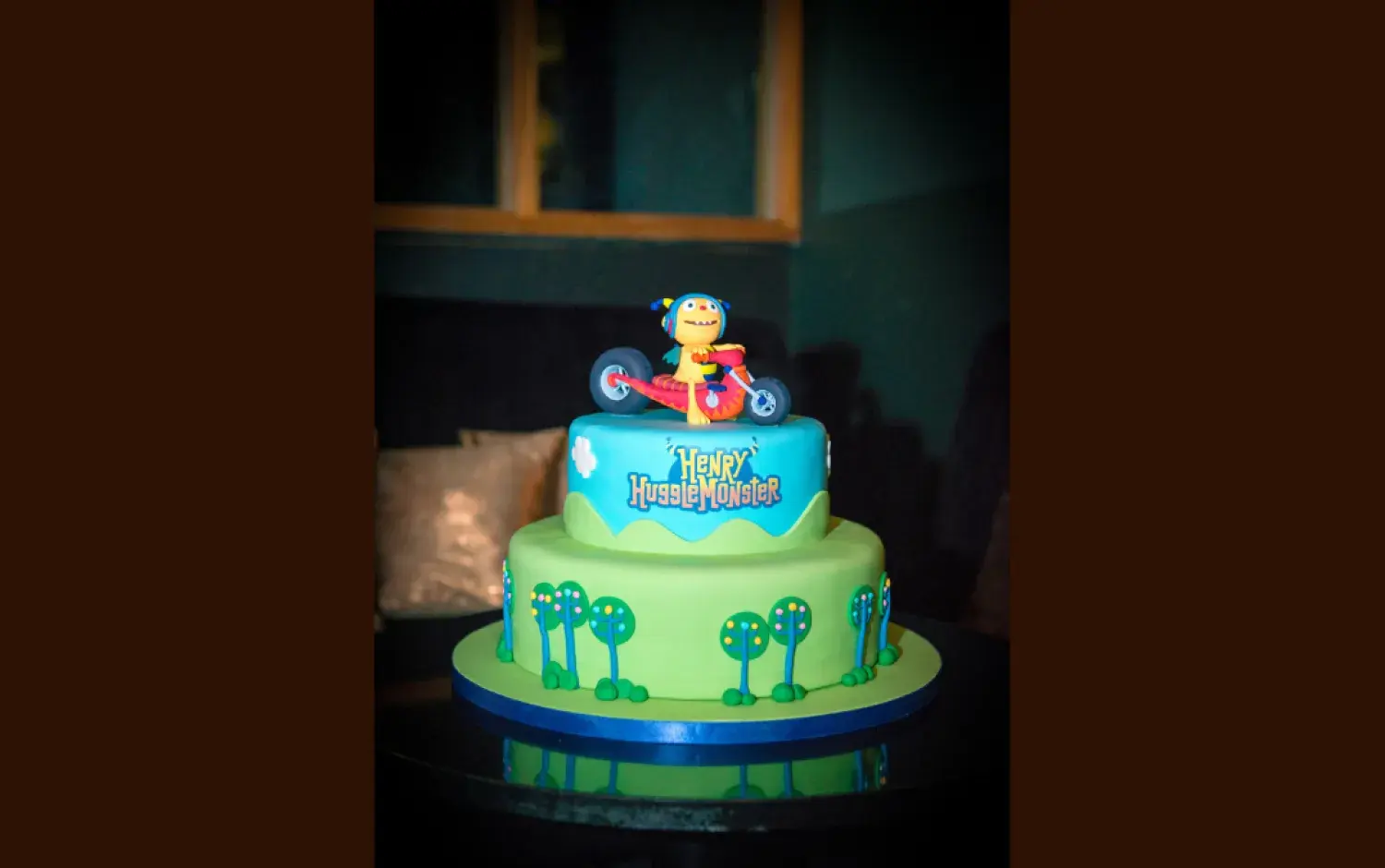 Two-tiered cake decorated with green and blue fondant and a figure of Henry Hugglemonster riding a tricycle on top.