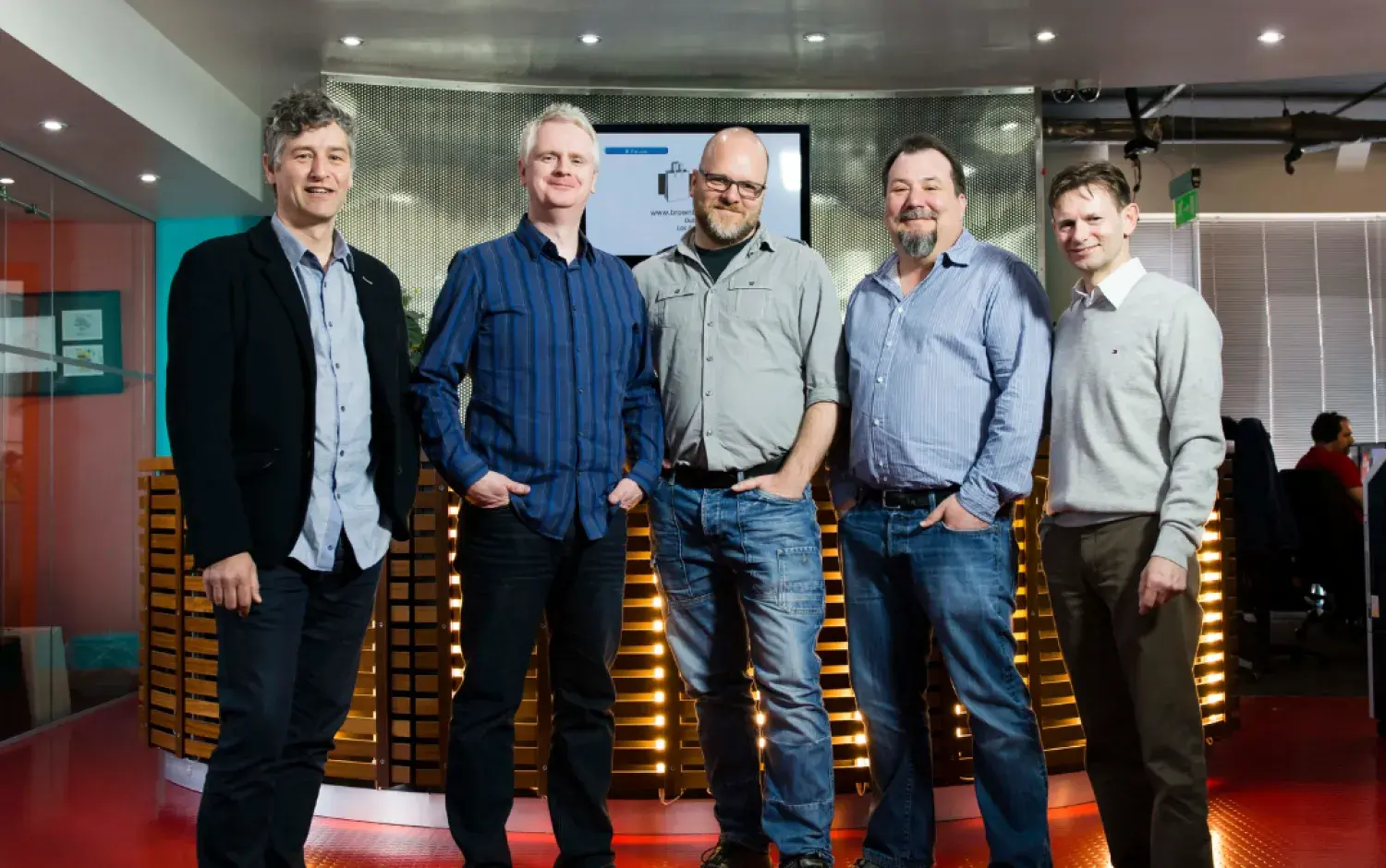 Five people standing together in a modern office space with a screen in the background.