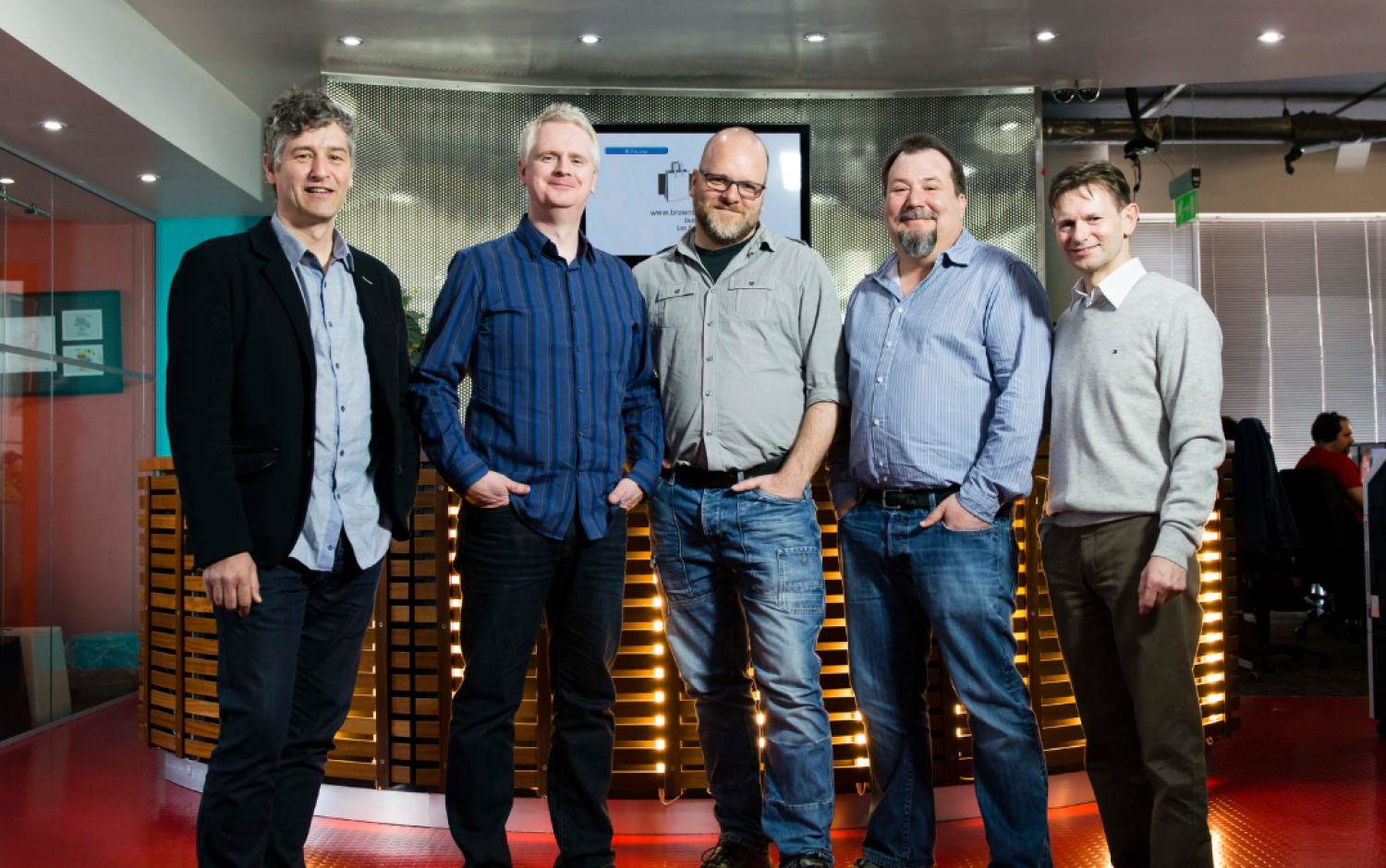 Five people standing together in a modern office space with a screen in the background.