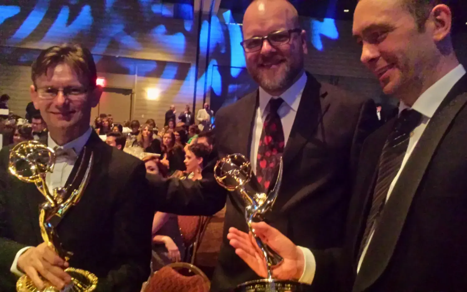 Three people in formal attire holding Emmy awards at a seated event with a crowd in the background.