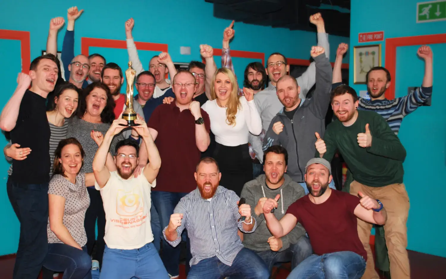 Large group celebrating indoors with raised arms, one person holding a gold trophy.