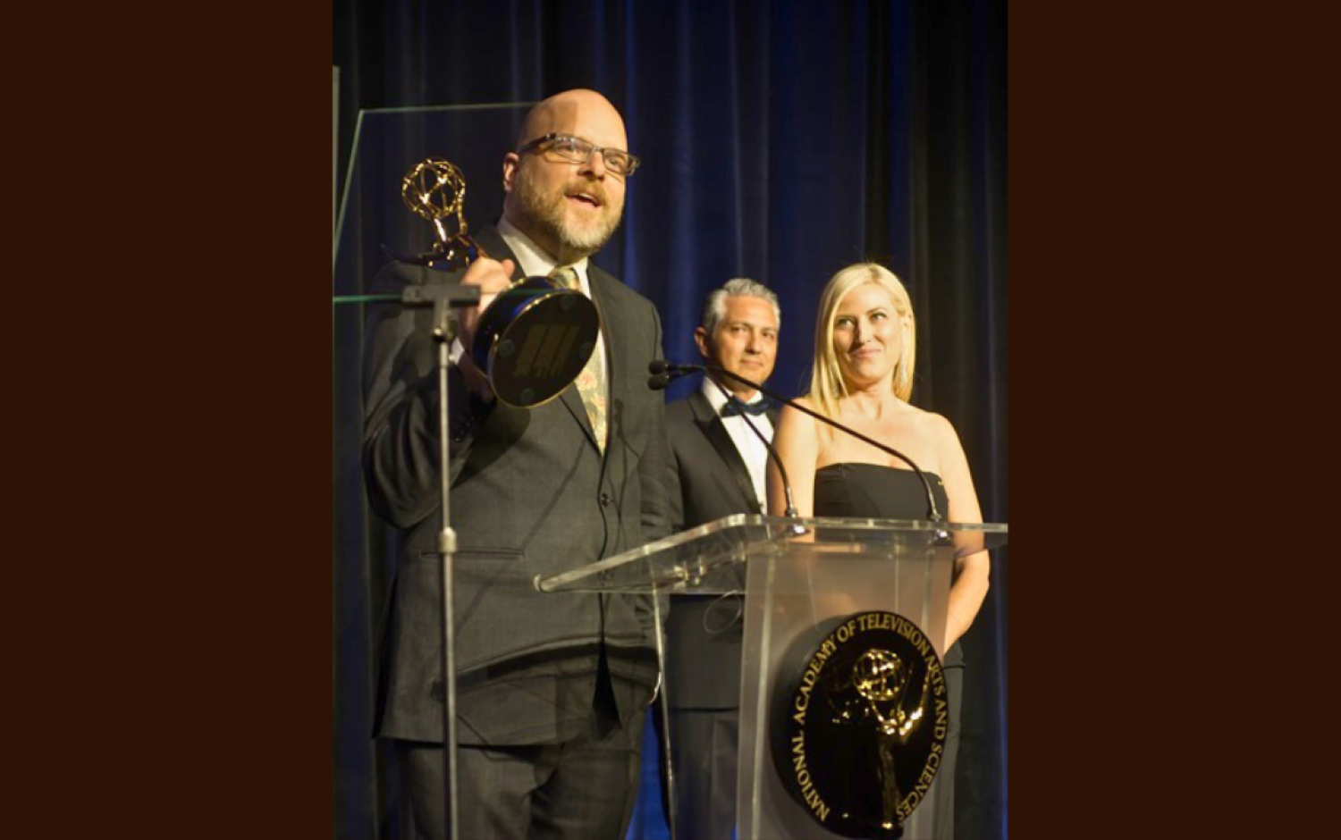 Person speaking at podium holding an Emmy Award during a formal ceremony.