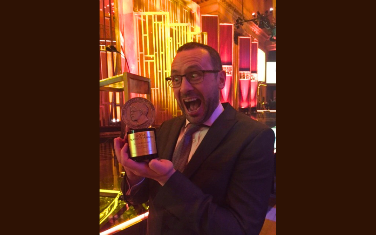 Person in a suit holding a Peabody Award trophy at an event with golden decor.