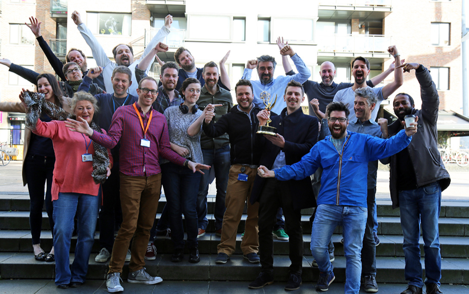 Large group outdoors posing with raised arms, one person holding an Emmy Award trophy.