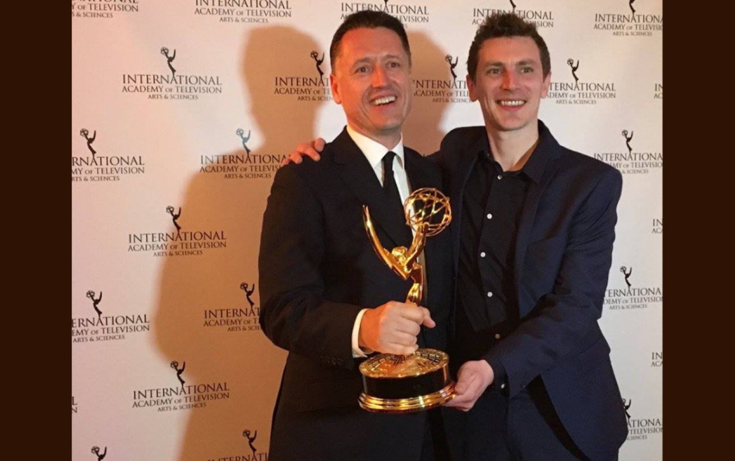 Close-up of two people holding an Emmy Award trophy against an International Academy of Television backdrop.