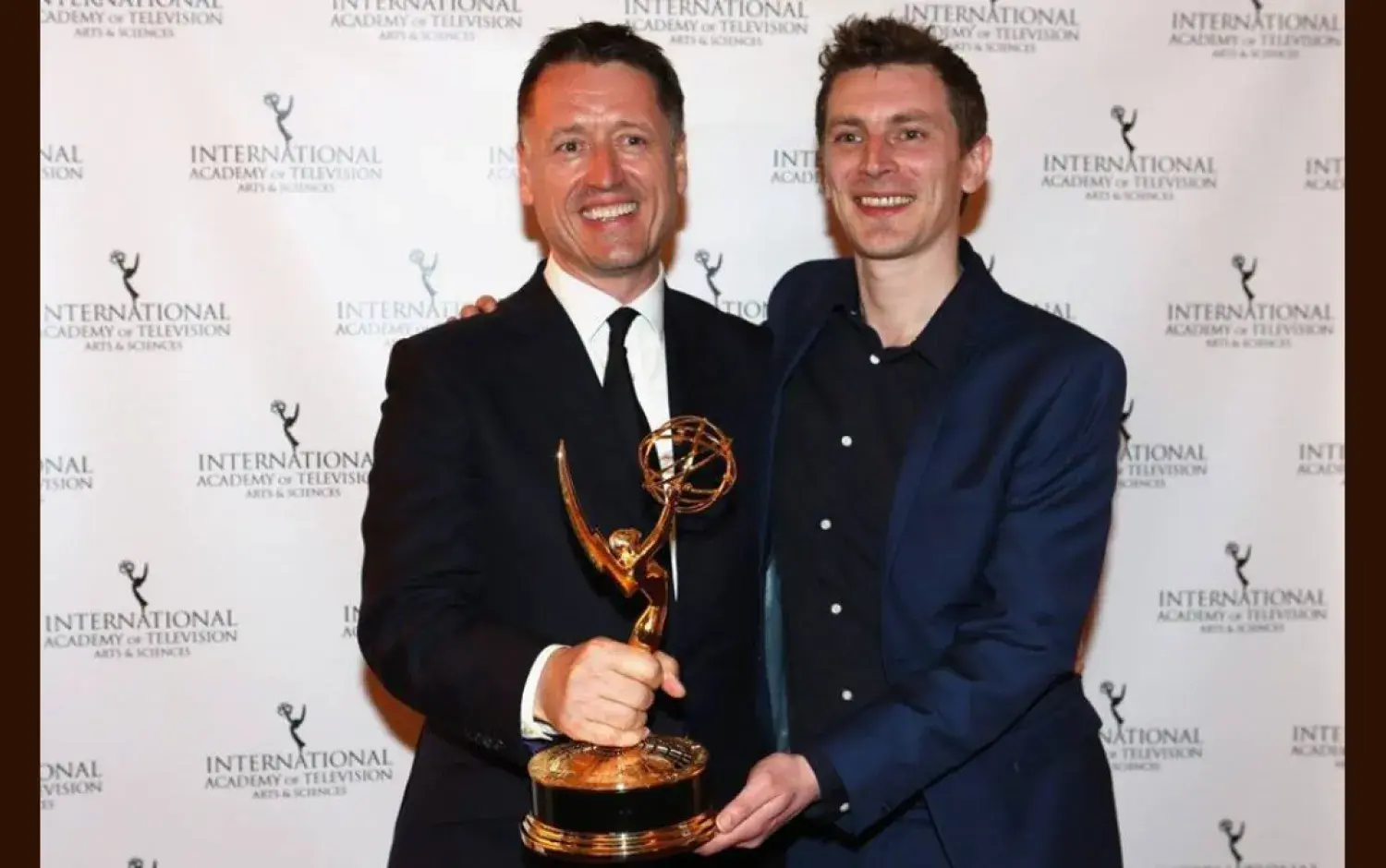 Two people holding an Emmy Award trophy in front of an International Academy of Television backdrop.