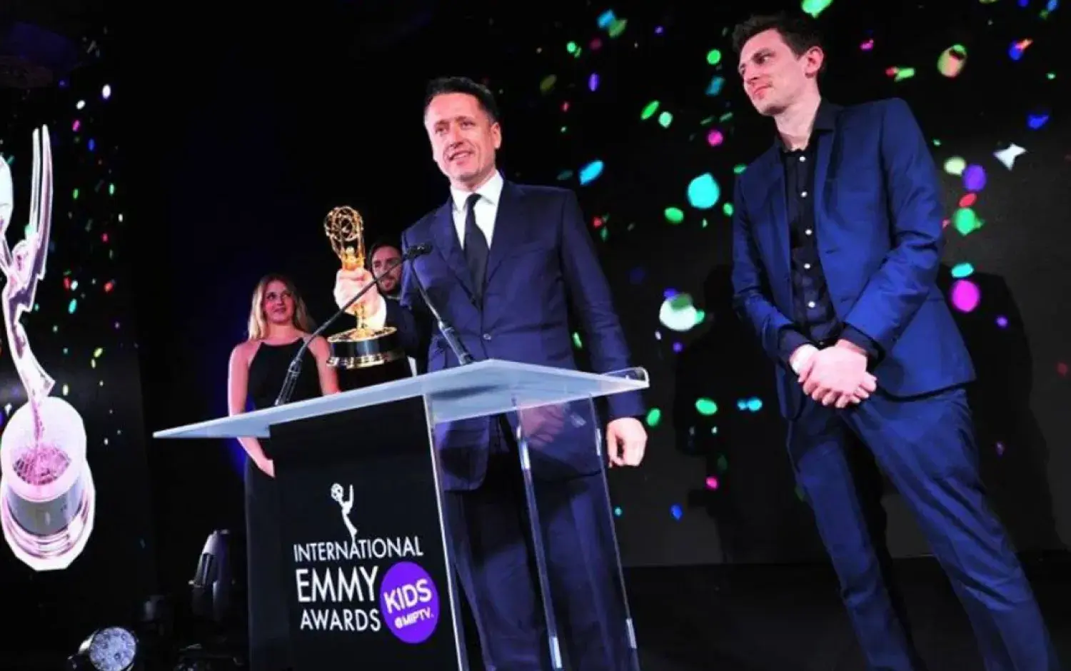 Two people standing at a podium with an Emmy Award trophy during International Emmy Kids Awards ceremony.