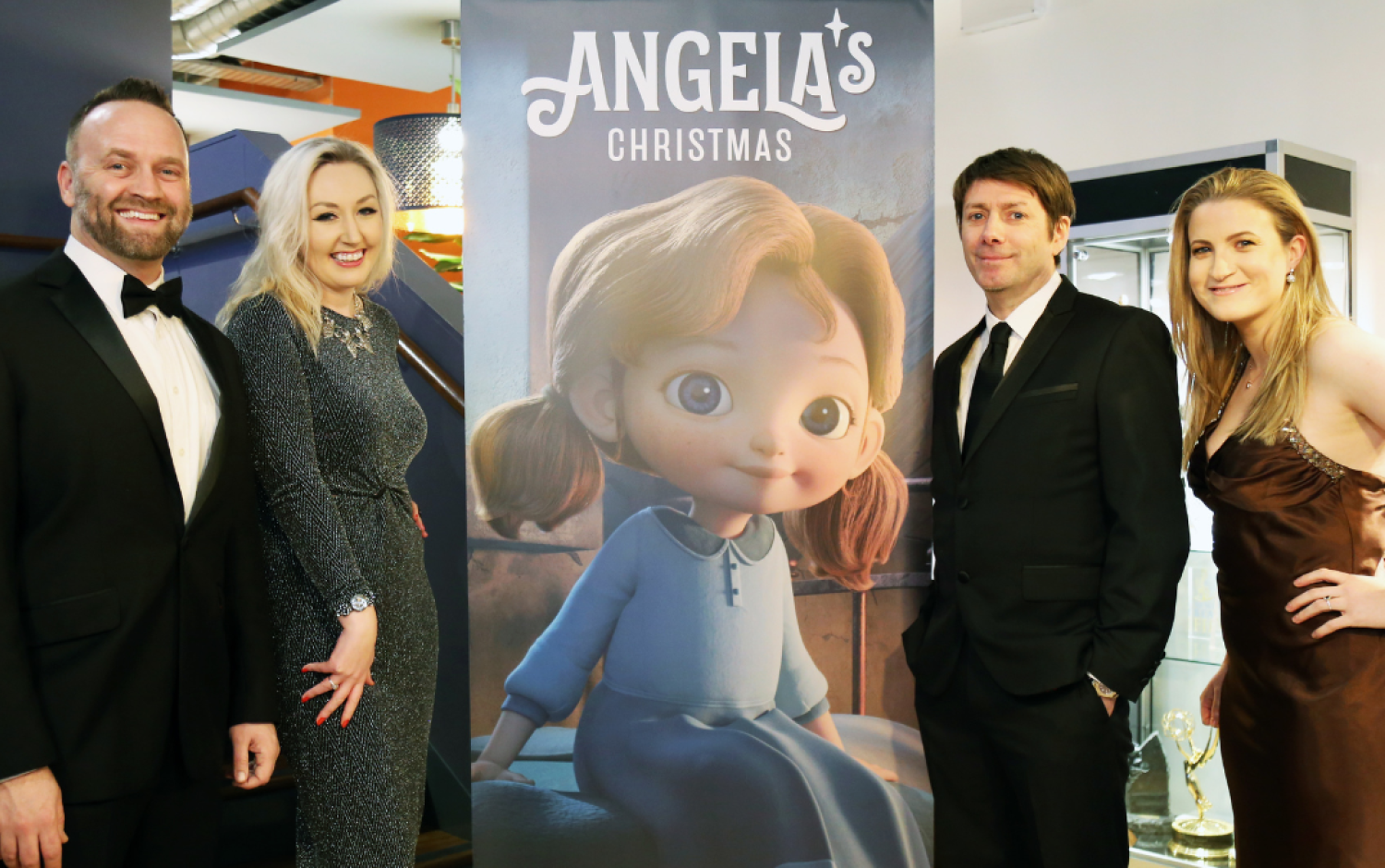 Group dressed in formal attire posing beside a large poster for “Angela’s Christmas.”