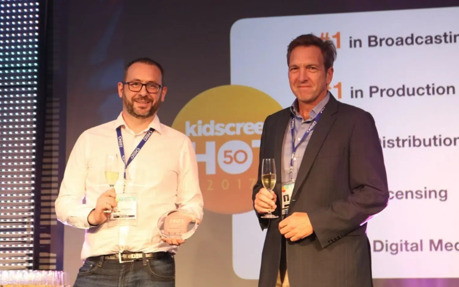 Two people on stage holding awards and champagne glasses with Kidscreen branding in the background.