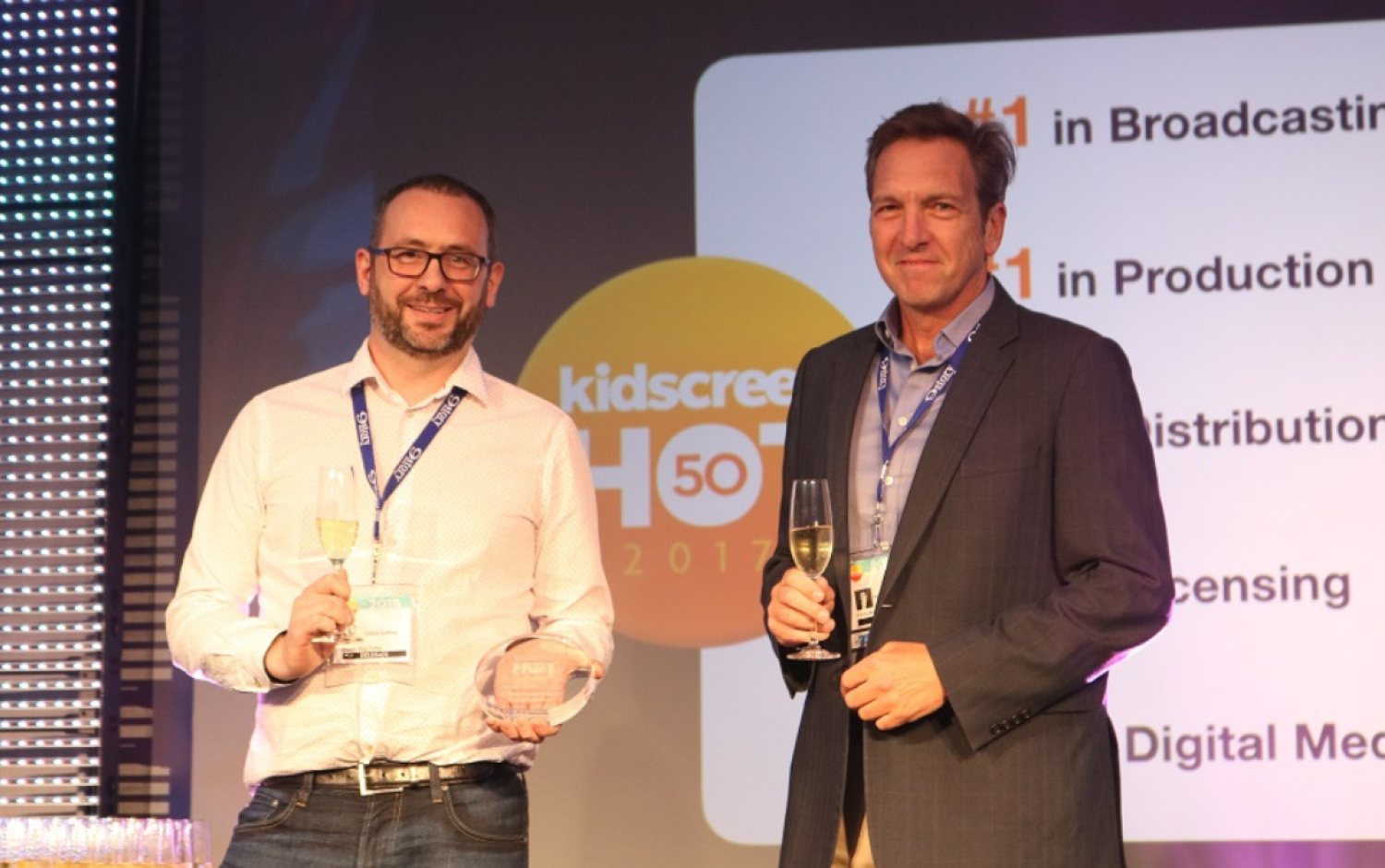 Two people on stage holding awards and champagne glasses with Kidscreen branding in the background.