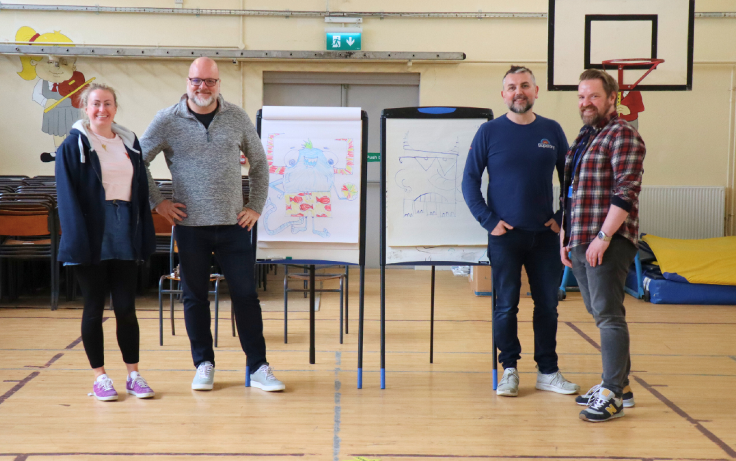 Four people standing in a gym beside flip charts displaying colorful drawings.